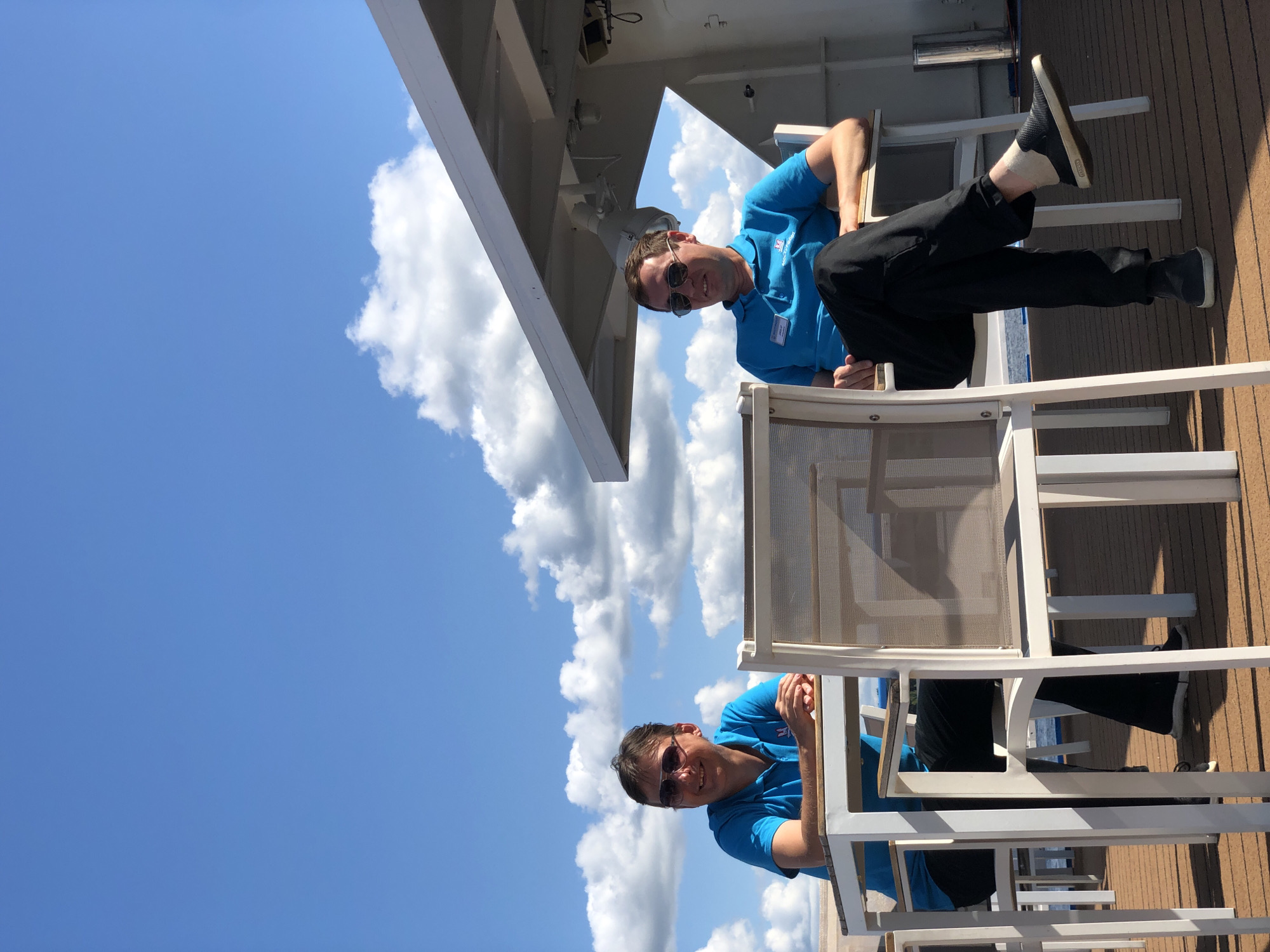 The image shows two men sitting at tables on an outdoor deck. Both men are wearing blue polo shirts and sunglasses. The man on the left is looking to his left and smiling. The man on the right is looking forward. There are chairs and tables around them, and a clear blue sky with white clouds is visible in the background. A portion of a white structure, possibly a canopy or roof, is visible above the man on the right. The deck appears to be made of wood. There is no indication that this scene is located in the city of Myshkin, Russia.