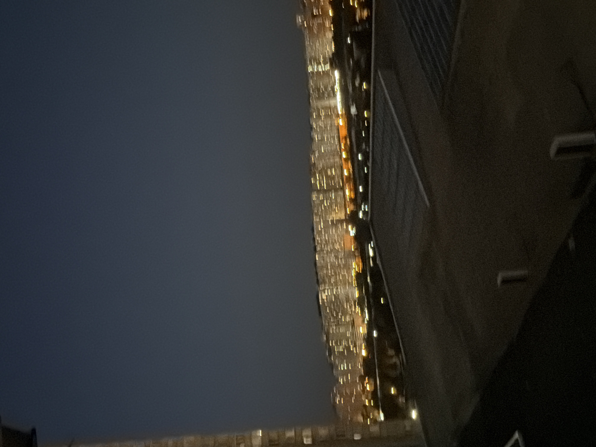 The image shows a blurred view of a city skyline at dusk or night, with numerous illuminated apartment buildings in the distance. The foreground features the roof of a building, with some dark structures and indistinct shapes. The sky is a dark blue-grey, and the overall impression is one of urban sprawl at night. There is no specific indication of the city being Kyiv, Ukraine, within the image itself.