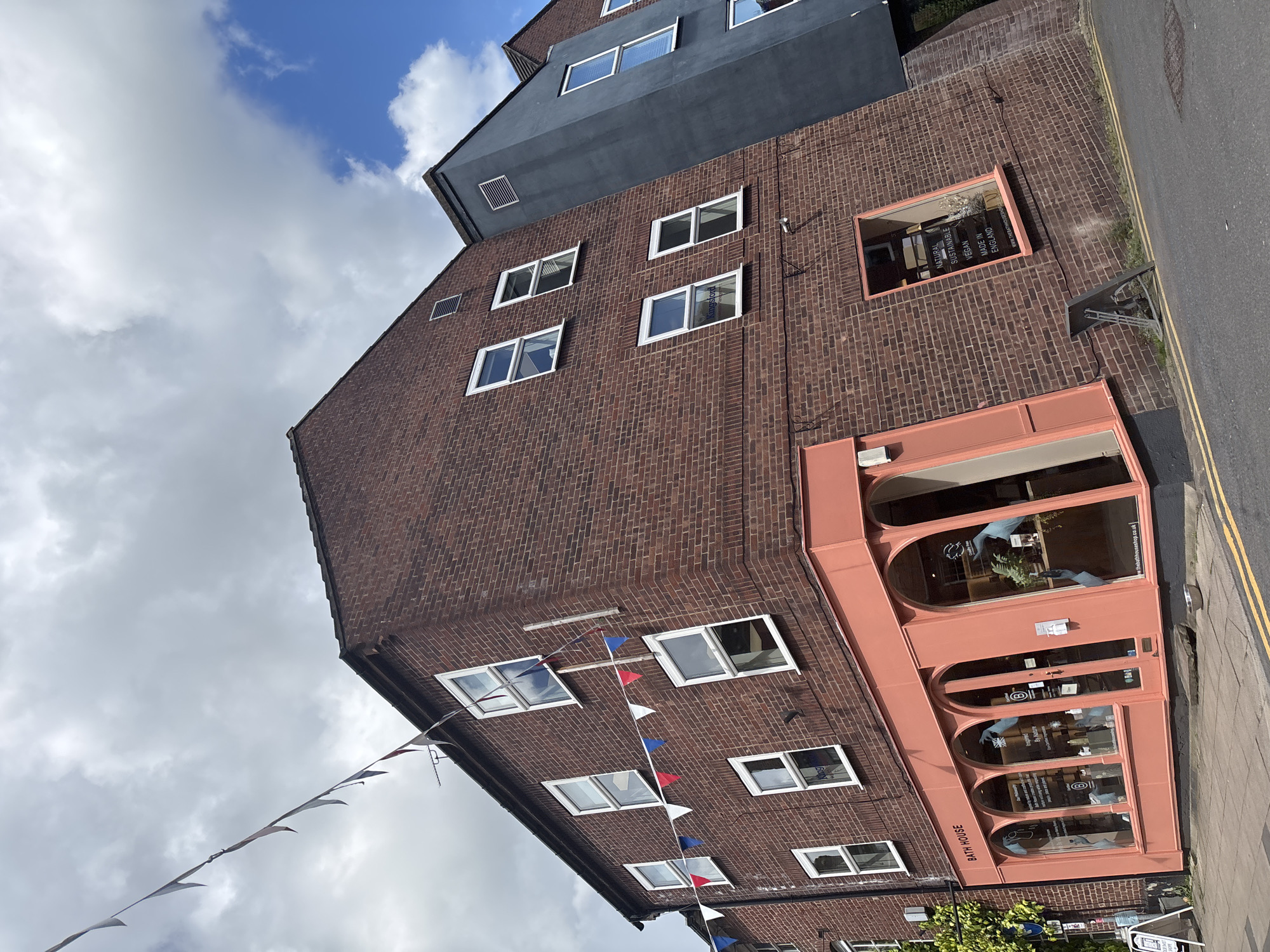 The image shows a brick building in Knutsford, United Kingdom. The building is multi-story, with a dark gray addition on top. It has numerous white-framed windows. The front of the building features a pink storefront with arched windows, labeled "BATH HOUSE." A string of red, white, and blue pennant banners is draped across the facade. A narrow sidewalk with a yellow line runs along the side of the building. The sky is partly cloudy.