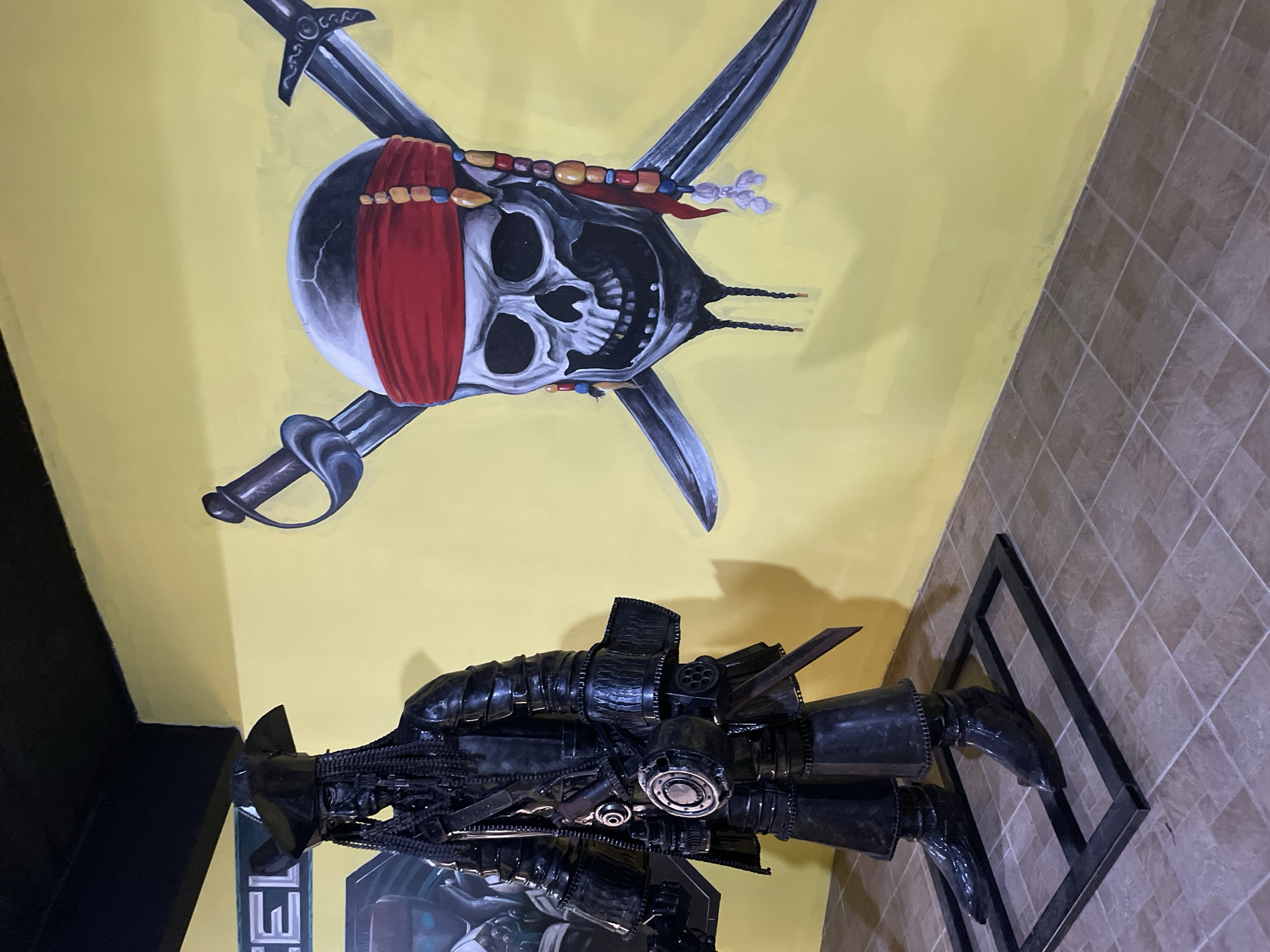 An outdoor scene at the pirate-themed attraction "Pirate Paradise" in Na Chom Thian, Thailand. A life-sized, dark-colored, metallic statue of Captain Jack Sparrow, depicted in his iconic pirate attire, stands on a black metal display frame. To the right of the statue, a large mural on a yellow wall features a pirate skull and crossbones with crossed swords and a red bandana. The ground is tiled with brown and beige squares.