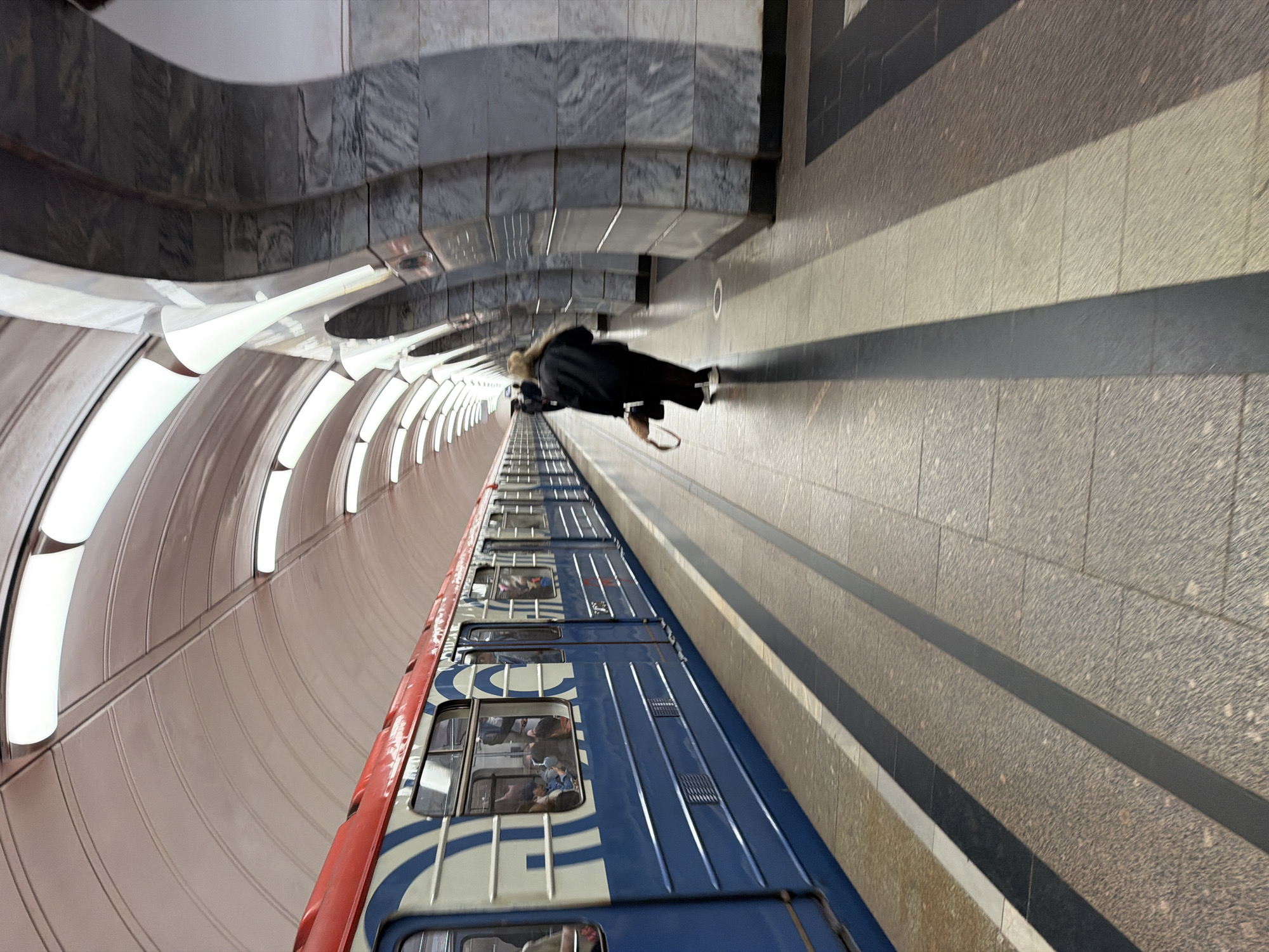 A long, blue and red subway train is stopped at a station platform. The train has white circular designs on its side. Inside the train, passengers are visible through the windows. A woman in a black coat and white sneakers is walking away from the train on the platform, carrying a brown bag. The subway station has a high, arched ceiling with integrated lighting. The platform is paved with granite tiles in various shades of gray and brown, with black safety lines. The station architecture features large, arched stone pillars. The image is taken from a slightly low angle, looking down the length of the platform.