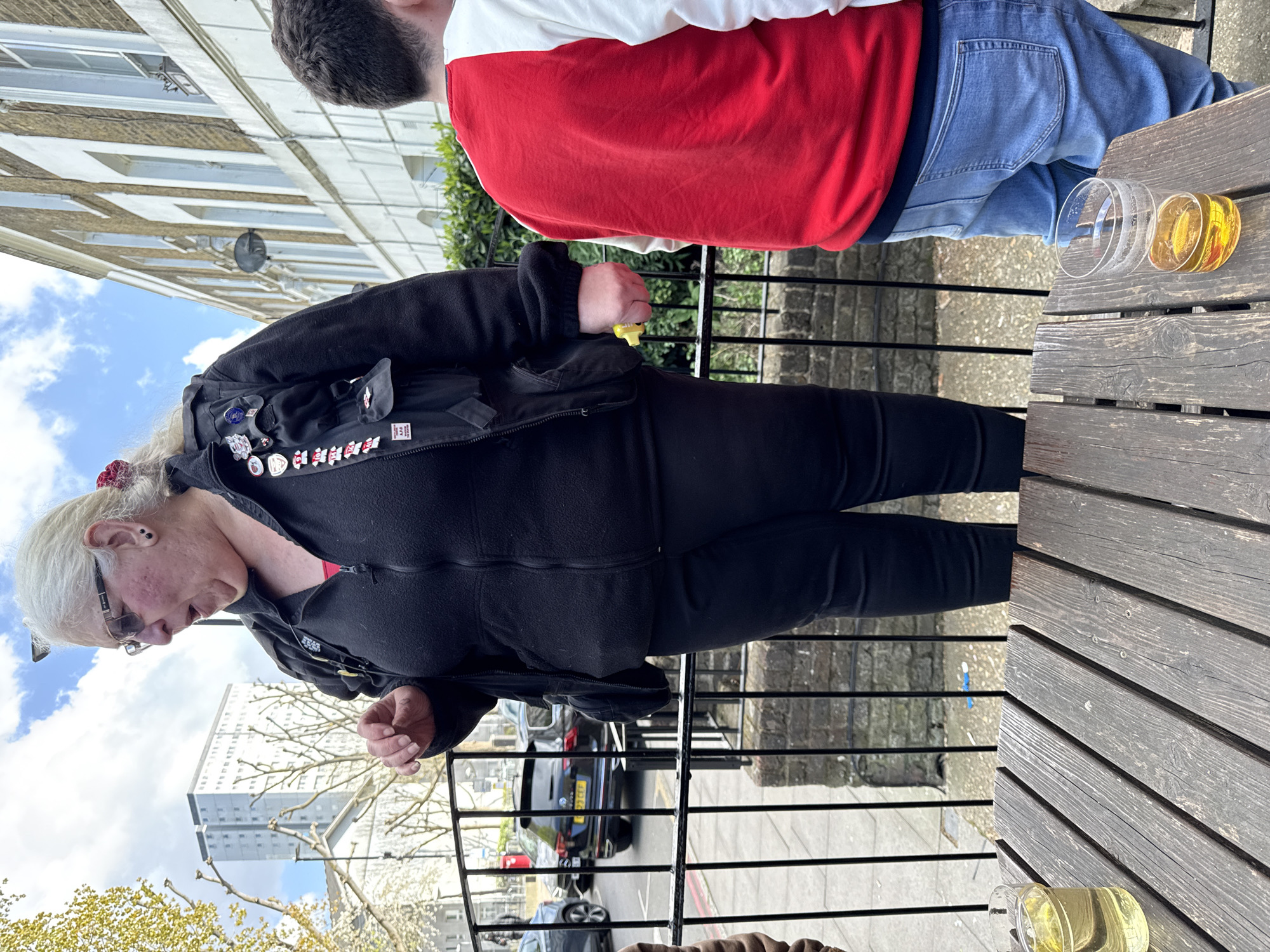 The image shows two people at an outdoor table. One person, with white hair and glasses, wears a black jacket with pins and black leggings. They appear to be looking down at something. The second person, partially visible, wears a white and red top with blue jeans. Two glasses of amber liquid sit on the wooden table. A black iron fence runs between the table and the street. In the background are cars, buildings, and trees under a blue sky with clouds. Based on the building architecture, the scene is potentially located in London, United Kingdom.
