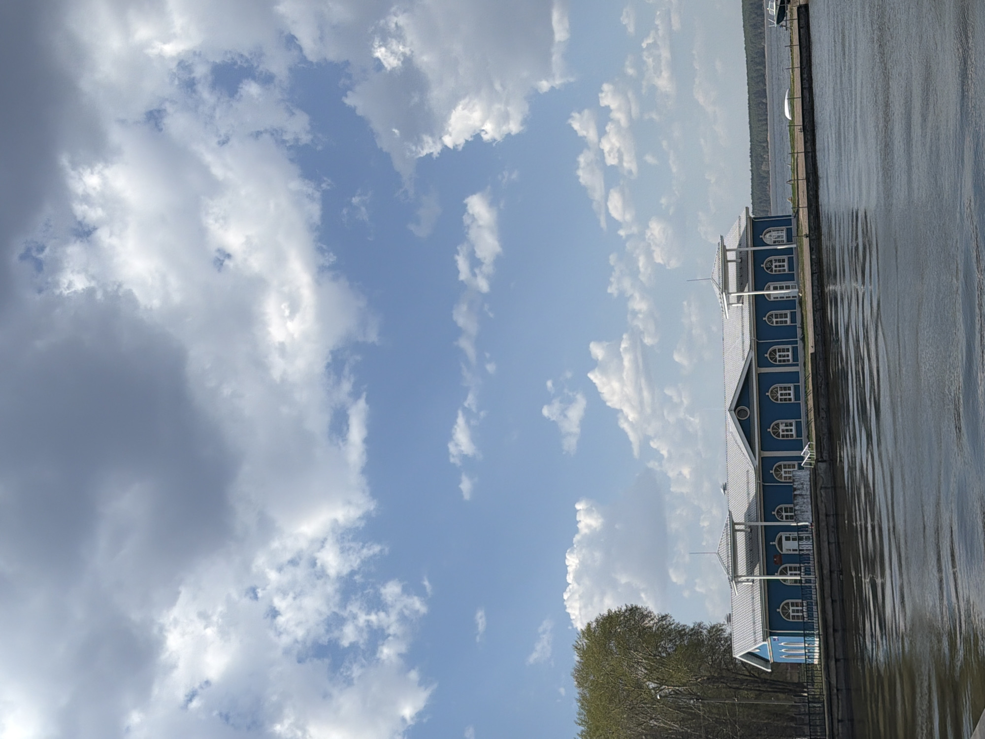 A daytime, rotated outdoor shot shows a building and water, with clouds occupying most of the sky. The building is blue with white-framed windows. It has a light gray roof, and a flagpole on top. Trees are visible at the building's base. The building is situated beside a body of water, and the water's surface is reflective. Above the building and water, a sky is full of white and gray clouds with patches of blue sky visible between. The location is likely in Votkinsk, Russia, given the prompt.