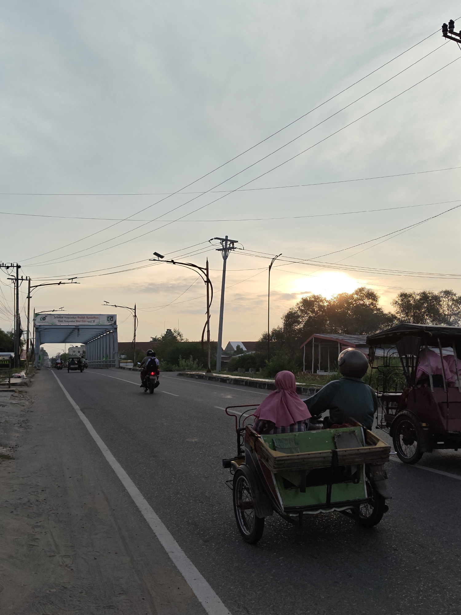 The photograph captures a street scene, likely during the early evening given the position of the sun low on the horizon. The location is suggested to be Lhokseumawe, Indonesia.
In the foreground, there are two "becak" or trishaws. One in front is green and the other is maroon. The photographer is also riding on a trishaw. The green one is carrying a passenger who is wearing a pink headscarf, and the driver of the trishaw is wearing a helmet. Further down the road, a motorcycle is also headed in the same direction as the trishaws.
The road is a two-lane paved highway with a white line marking the edge of the lane. A bridge with an archway featuring what appears to be signage can be seen farther down the road. The text "Selamat Menjalankan Ibadah Ramadhan Dan Menyambut Idul Fitri 447 31" is visible. Electrical poles and wires stretch overhead, intersecting the sky.
The sky is partially cloudy, and the sun is positioned behind some trees, giving a warm glow to the scene. Vegetation can be seen on either side of the road, and there are some buildings in the background.