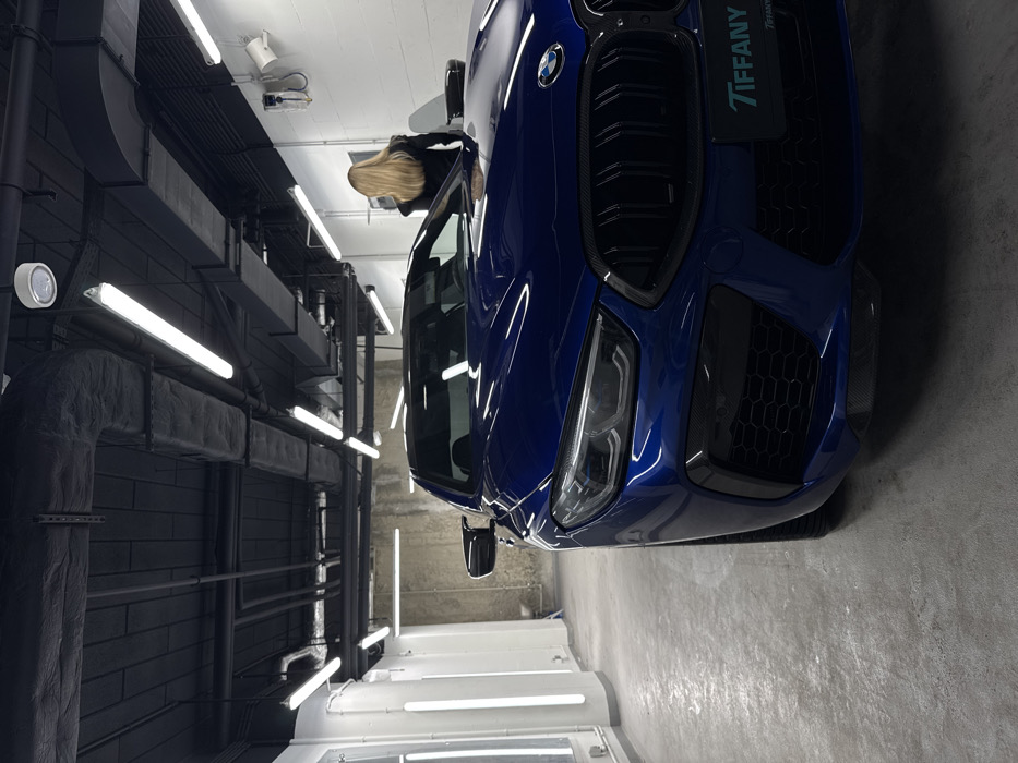 The image displays the front left three-quarter view of a dark blue BMW M8 Series coupe, stationary within what appears to be a professional car detailing or service bay in Warsaw, Poland. The location is Tiffany Car Detailing & PPF at Kolejowa 49, 01-210 Warsaw, as indicated by a "TIFFANY" plate on the car's front bumper.
The environment features a light gray, reflective concrete floor and bright, even illumination from multiple suspended linear fluorescent light fixtures. The ceiling is black, exposing ducts, pipes, and electrical conduits. Walls consist of white painted surfaces on the right and a raw, textured concrete wall further back on the left, where vertical fluorescent tubes are mounted.
A woman with long blonde hair, dressed in a dark, long-sleeved top or jacket, stands behind the car on the right side of the frame, facing away from the camera and towards the rear of the vehicle. Her posture suggests observation. On the white wall directly behind her, a wall-mounted electrical unit or charging station is visible. The BMW exhibits a glossy finish, detailed with black kidney grilles, intricate headlight design, and honeycomb mesh inserts in the lower bumper, consistent with a premium sports vehicle maintained in a specialized facility.