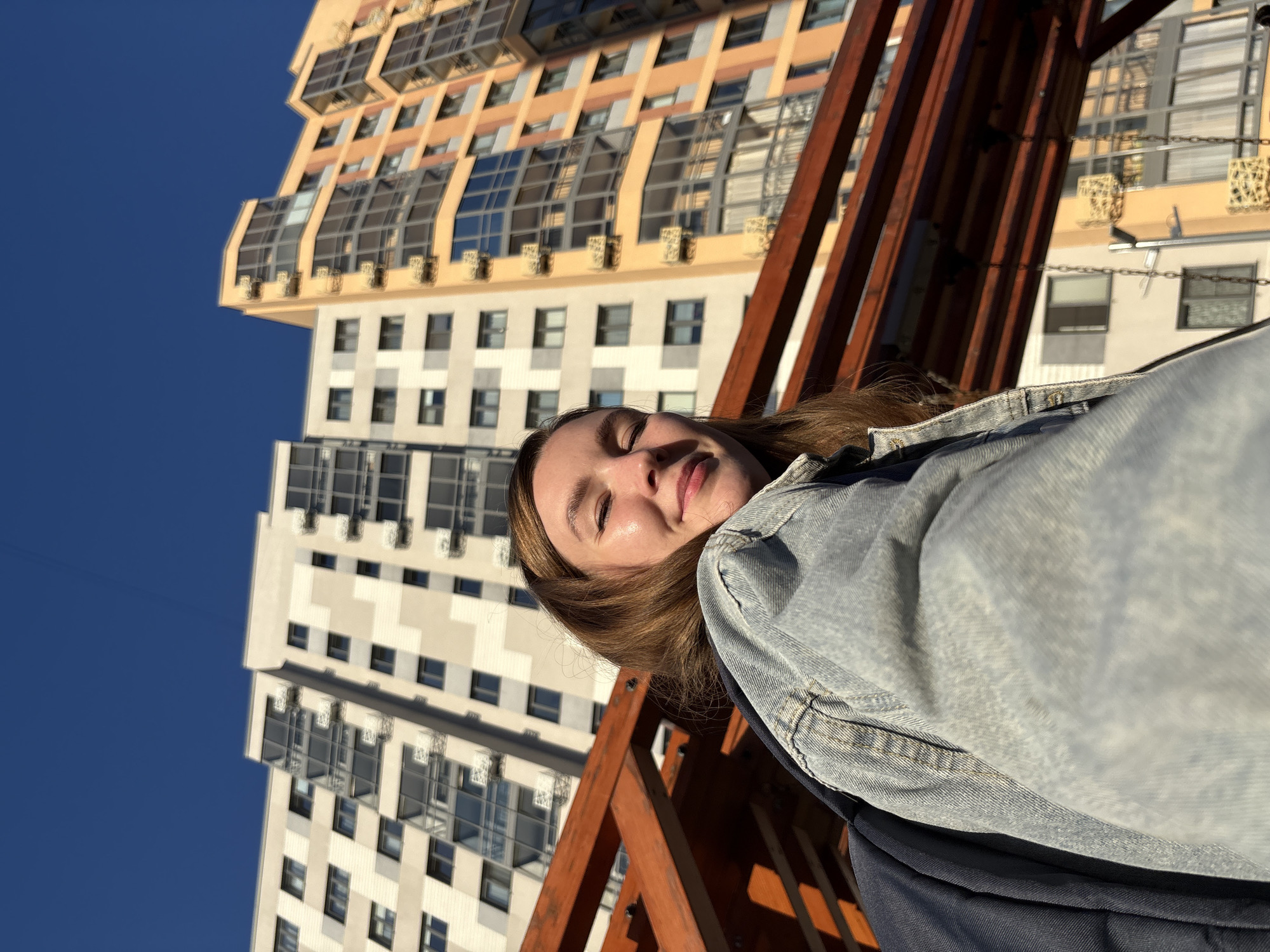 A young woman with brown hair and closed eyes smiles into the camera, her face angled upwards. She wears a light blue denim jacket and a dark blue backpack. Behind her, two tall apartment buildings rise against a clear blue sky. The building on the left is primarily white and grey with dark windows, while the building on the right is a vibrant orange with a curved facade and dark tinted windows. In the foreground, a wooden structure, likely part of a playground or outdoor seating area, casts a shadow. The image appears to be taken outdoors during the daytime, with sunlight illuminating the woman's face. The buildings suggest an urban environment, and the architectural style is consistent with modern residential complexes. This scene is likely located in Izhevsk, Russia, given the context provided.