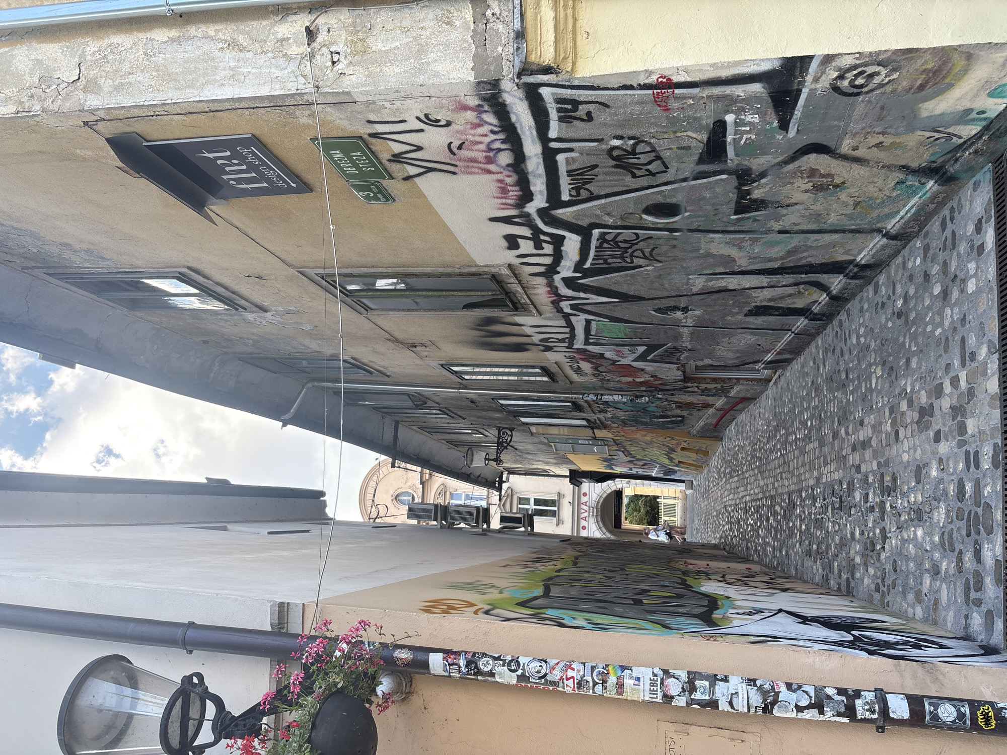 The image depicts a narrow, graffiti-covered alleyway. The buildings lining the alley have a weathered appearance, with cracked concrete and peeling paint. The walls are extensively covered in various forms of graffiti, ranging from large, bold lettering to more intricate designs. A cobblestone pavement runs along the right side of the alley.
On the left side of the alley, a sign is visible that reads "Flirt design shop" with an arrow pointing to the right. Another green street sign is also visible, with text that appears to be in a non-Latin script, possibly Cyrillic or Greek, with the number "3" below it.
Further down the alley, an archway leads to a brighter area where a few people are visible. One person is seated on a white object, and another appears to be walking. The sky visible in the background suggests it is daytime, with some clouds present. The overall impression of the alley is one of urban grit and artistic expression. A lamppost with pink flowers is visible in the foreground on the left. The scene evokes a sense of mystery and exploration, with the graffiti adding a layer of vibrant energy to the otherwise aged architecture.