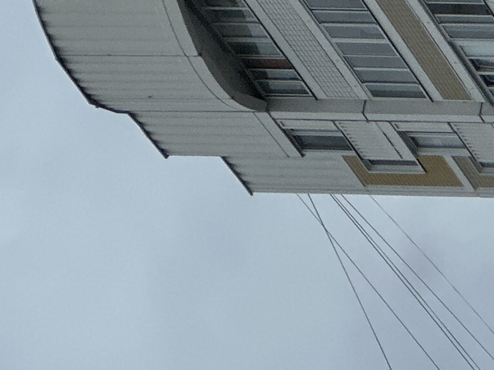 The image shows a close-up of a residential building in Podolsk, Russia. The building appears to be a multi-story apartment complex with a modern facade. The sky is overcast and gray, suggesting cloudy weather. Several thin, dark electrical or telephone wires stretch diagonally across the left side of the frame, intersecting with the building. The building itself features a curved section at the top and rows of windows, some with balconies. The architectural style suggests a post-Soviet era construction. There are no people visible in the frame. The scene is an outdoor urban environment, focusing on the side of a building and the sky.