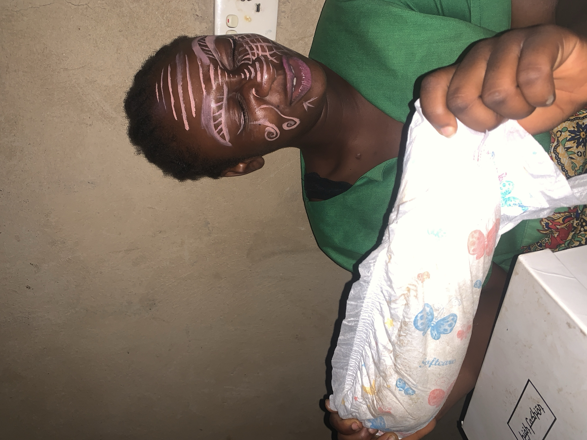 A young Black woman with face paint is holding a diaper. She is wearing a green shirt. The background is a plain wall. The scene is indoors. The woman's face paint includes pink lines on her forehead and around her eyes and nose, as well as swirls on her cheeks. The diaper is white with colorful butterfly and flower prints. There is a white box in the foreground. The woman's eyes are closed, and she appears to be smiling or laughing.
