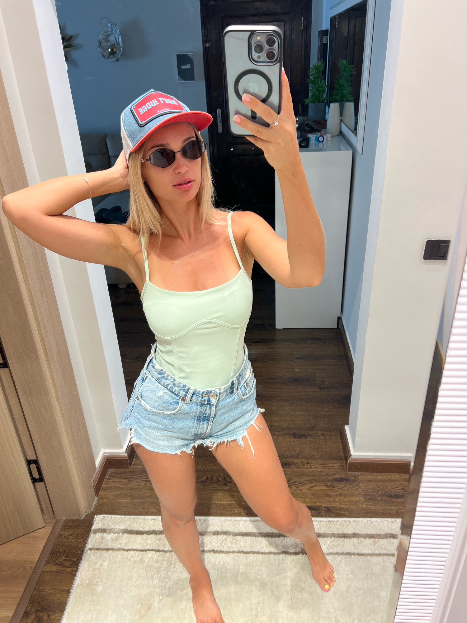 A woman stands in what appears to be a residential interior, possibly a bedroom or hallway, in Sharm El Sheikh, Egypt. She is taking a mirror selfie. She has blonde hair and is wearing a light green tank top, distressed denim shorts, sunglasses, and a baseball cap. Her left arm is raised, and she is holding the back of her head. The background shows a mirror reflecting a dresser with decorative items, a doorway, and a wooden floor. The lighting suggests it is daytime.