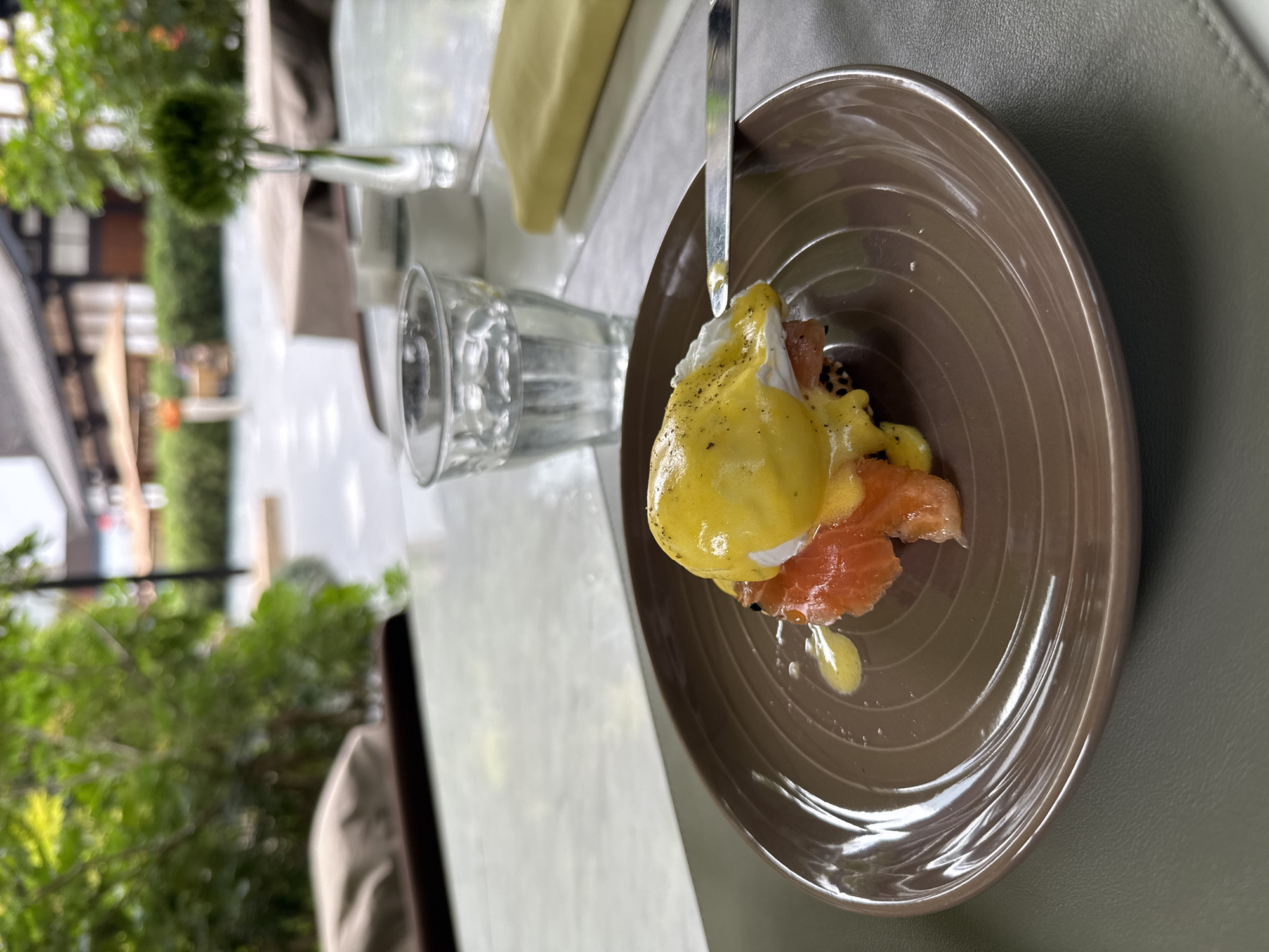 A plate of Eggs Benedict with salmon sits on a table with a glass of water and a napkin, in an outdoor setting that may be a patio. The dish consists of salmon, a poached egg, and hollandaise sauce. The photo was taken in the Jinjiang District, China.