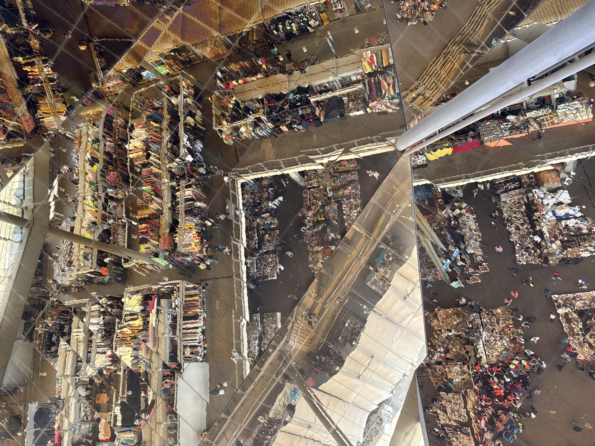 This image captures a high-angle view of a bustling indoor marketplace, viewed through a complex, multi-faceted mirrored ceiling. The scene below is filled with numerous people, appearing as small figures, actively browsing and shopping among a multitude of stalls.
**Subjects:** The primary subjects are the crowds of people populating the market floor, along with vast displays of goods. These goods consist predominantly of clothing, textiles, and various bric-a-brac, arranged on racks, tables, and in large piles directly on the floor. The architectural feature of a large, grey cylindrical column is visible, cutting through some of the mirrored reflections. The mirrored ceiling itself, composed of many reflective panels, is a key subject, distorting and multiplying the view of the market below.
**Activity or event taking place:** The image depicts a vibrant and busy commercial event, likely a flea market or a large retail market. People are engaged in typical shopping activities: walking through aisles, observing merchandise, and interacting with stalls. The atmosphere appears lively and crowded.
**Notable details:** The most distinctive feature is the highly reflective, possibly mirrored, ceiling. This ceiling creates a mesmerizing kaleidoscopic effect, fracturing and multiplying the market scene into numerous distorted reflections and geometric patterns. This design element disorients the viewer's perception of space, making the market appear vast and intricate. The overall lighting suggests an indoor setting, likely during the daytime, with a combination of natural and artificial light sources reflecting off the surfaces. The fragmented reflections offer glimpses of the market's structure, revealing distinct sections and pathways. Given the context of Barcelona, Spain, this could potentially be the Mercat dels Encants Vells, known for its iconic mirrored canopy.
**Visible text:** No clearly legible text is discernible within the image. There are a few small, indistinct signs, one green, but any text on them is too blurry to read.