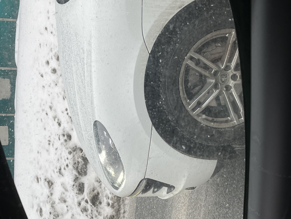 An image captured from within a vehicle shows the front left quarter of a white Porsche Macan SUV. The vehicle's white body panels, including the hood, fender, and bumper, are lightly dusted with ice pellets or melting snow. The left front headlight is visible and appears to be illuminated or reflecting light. The left front wheel features a silver multi-spoke alloy rim with a visible Porsche center cap and a brake caliper. The tire is a black winter type, heavily encrusted with snow or ice pellets. The ground directly beneath the vehicle is a dark, wet paved surface with remnants of slush. In the background, to the left and rear of the Porsche, lies a large mound of dirty, partially melted snow. Beyond this snowbank, a green fence or barrier with lighter patches is visible. The scene suggests cold, wintery conditions with light precipitation. This scene is located in the city of Solnechnogorsk, Russia.