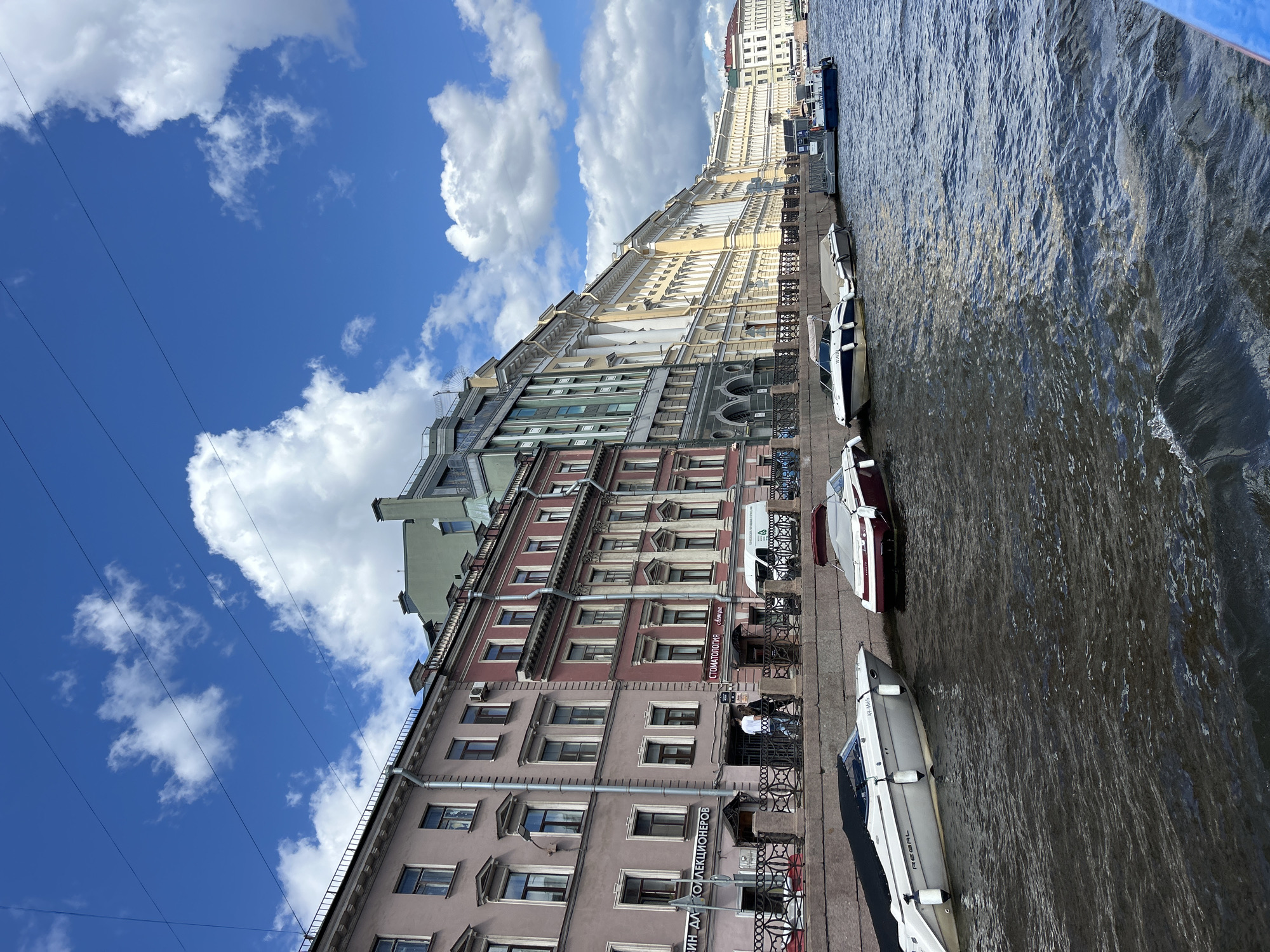 A canal-side view of historic buildings in an urban environment shows a line of boats docked along a stone embankment. The buildings are a mix of architectural styles and colors, with some featuring ornate facades and others appearing more modern. Several boats, ranging from small motorboats to larger vessels, are moored along the water. People are visible on the embankment, walking along the path. The sky above is blue with scattered white clouds. The water in the canal is dark and rippled.