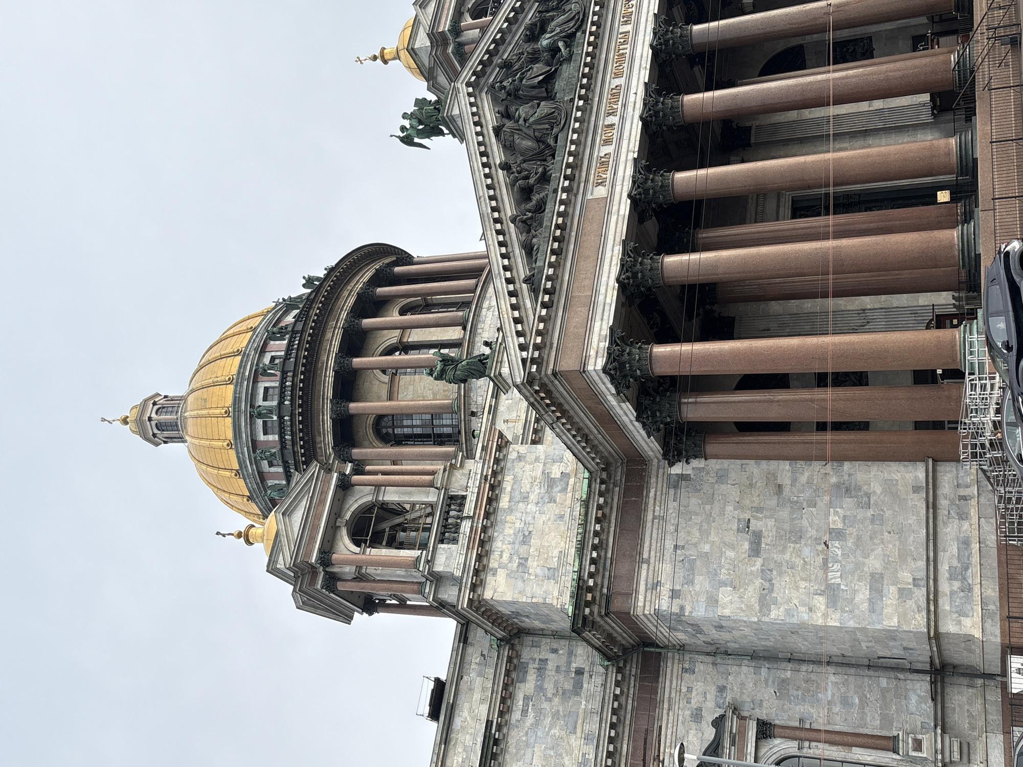 The image depicts the facade of St. Isaac's Cathedral in Saint Petersburg, Russia, viewed from a low angle, looking upwards. The grand building features a prominent golden dome, ornate architectural details, and large columns. The sky is overcast and a pale gray. In the foreground, a black car is parked behind a fence. The style of the architecture suggests it is a significant religious or historical landmark.