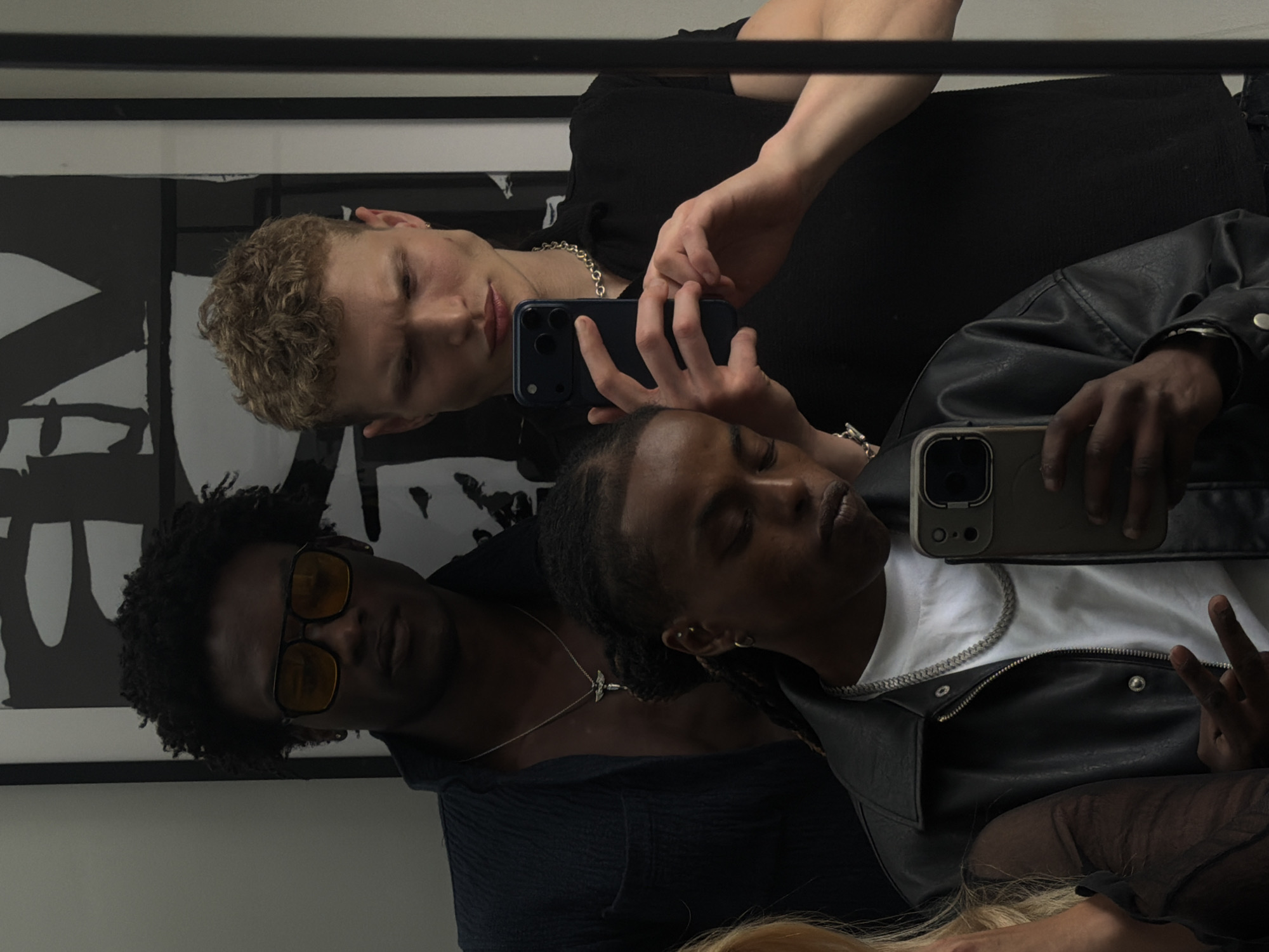 The image depicts a mirror selfie featuring three people, taken in an indoor setting in London, United Kingdom. The photograph is oriented vertically, showing the subjects as if they are standing or leaning within the reflection.
In the foreground, on the left, a Black man with short, dark curly hair and yellow-tinted sunglasses wears a dark-colored top and a silver necklace with a cross pendant. To his right, a second person, also Black, with dreadlocks, is visible. They wear a white t-shirt underneath a black leather jacket, accessorized with a silver chain necklace. This individual holds a light-colored smartphone and is seen making a peace sign with their left hand. Behind them, partially obscured, is a third person with short, curly blonde hair, wearing a black ribbed top and a prominent silver chain necklace. This person is holding a dark-colored smartphone, seemingly taking the photo, and is looking downwards. A small section of blonde hair, possibly belonging to a fourth person or an extended part of the third person's hair, is visible at the bottom right.
The background reflects a framed piece of abstract black and white art, suggesting a modern interior. The subjects appear to be casually dressed, and the activity is a shared mirror selfie. The lighting is soft and even, typical of an indoor environment. No specific text is visible within the image.