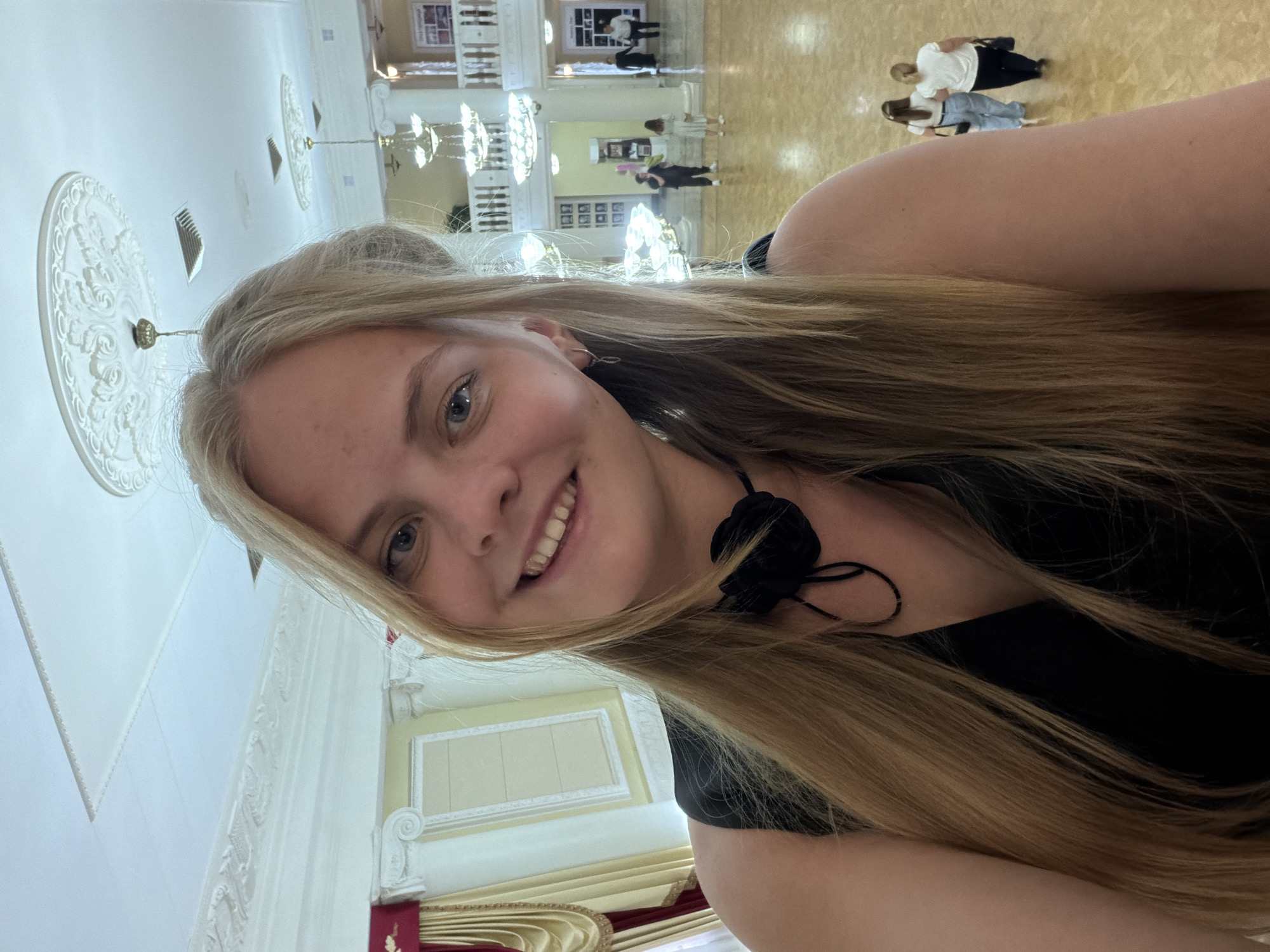 A young, fair-skinned woman with long blonde hair smiles into the camera. She is wearing a black top with a black flower detail near her neck. The background shows an ornate interior, likely a ballroom or event space, with high ceilings, decorative plasterwork, and chandeliers. In the distance, on a balcony, several people are visible. Based on visual cues, the location is likely the Izhevsk State Agricultural Academy's Assembly Hall in Izhevsk, Russia, known for its architectural details and event spaces.