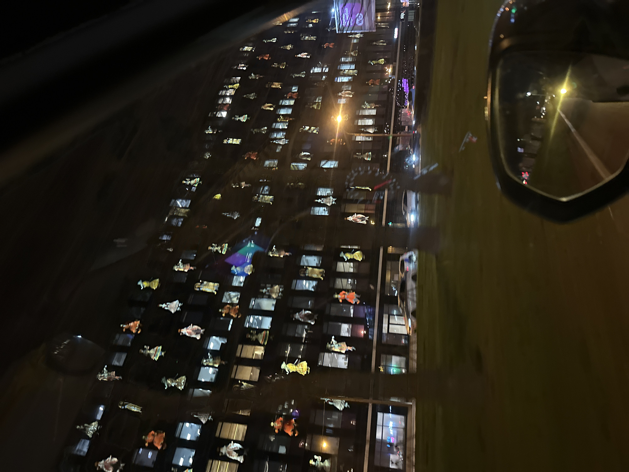 The image shows a building with windows decorated with figures. It appears to be taken from inside a car at night. The side mirror of the car is visible, reflecting streetlights and the road. The building with decorated windows seems to be located in Saint Petersburg, Russia. The figures decorating the windows are likely related to a holiday or event.