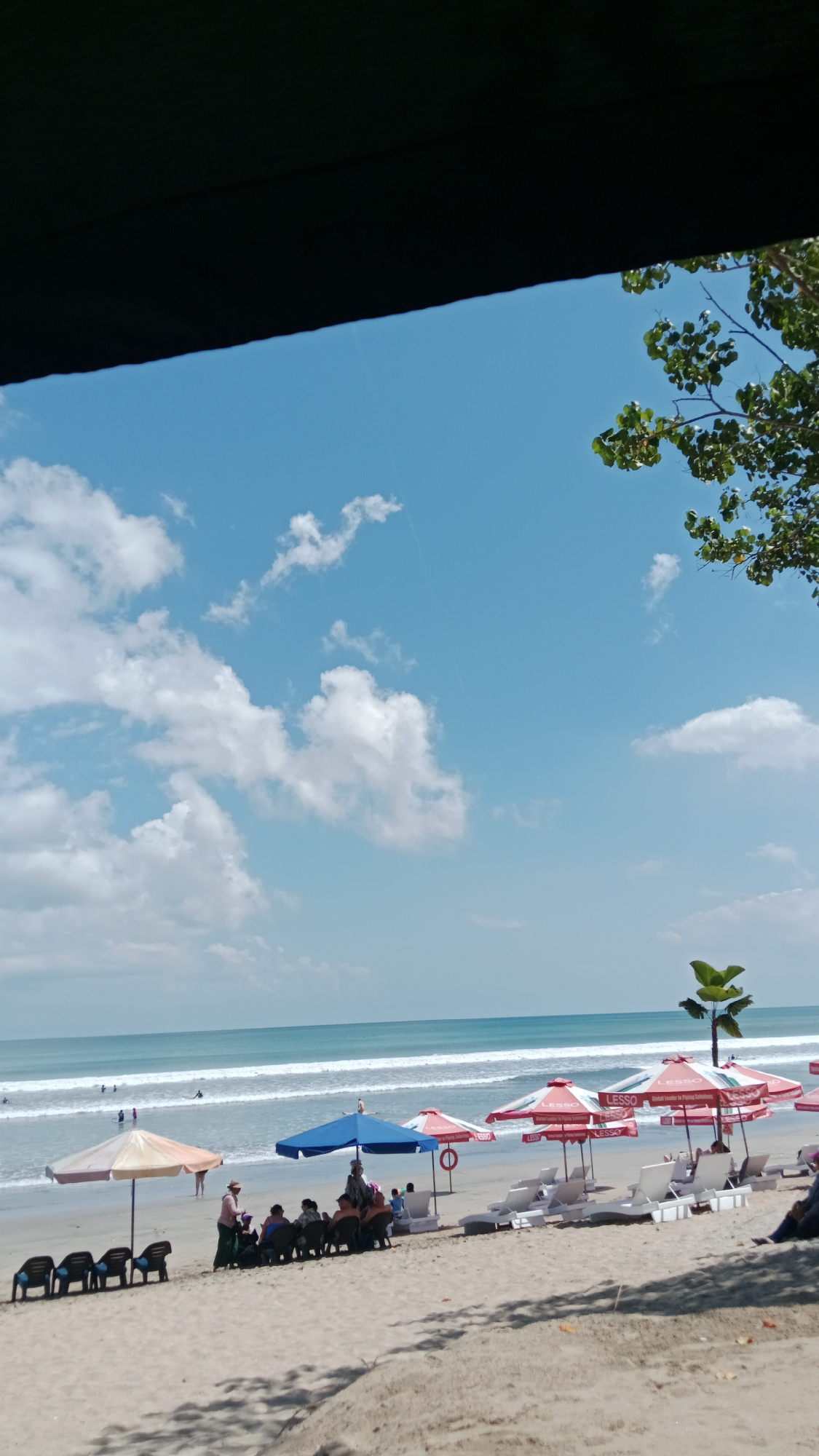 This is a photograph of Kuta Beach in Kuta, Indonesia. The image captures a sunny day with a clear blue sky and scattered white clouds. In the foreground, a sandy beach is populated with lounge chairs and umbrellas. Several people are seated under a blue umbrella, and others are visible further down the beach near the water. The ocean has gentle waves, and a few individuals can be seen swimming or enjoying the water. The overall scene depicts a typical relaxed atmosphere at a popular beach destination.