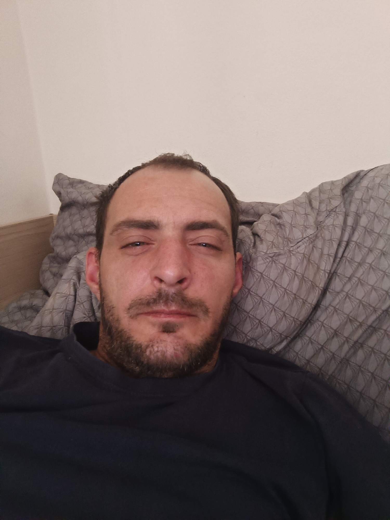 A man with dark hair, a beard, and a goatee is lying down, possibly in a bed, as indicated by the presence of a pillow and bedding with a diamond pattern. He is wearing a dark blue t-shirt. The background appears to be a plain wall, and a wooden headboard is visible on the left side of the image. The image is a close-up, selfie-style photograph. The specific location in Milovice, Czechia cannot be determined from the visual information provided.