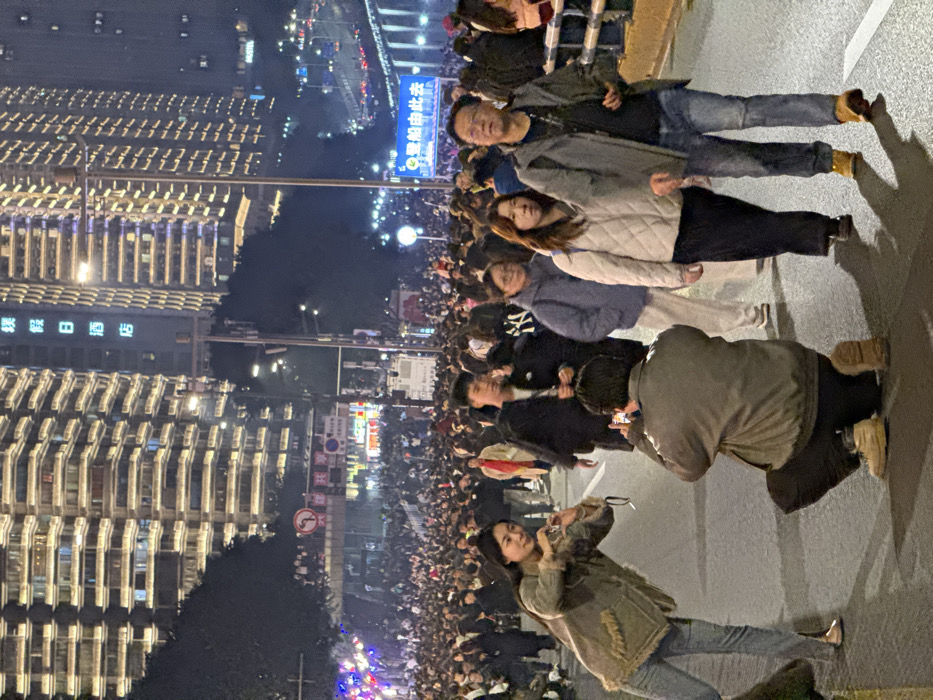 A dense nighttime street scene is depicted in Yuzhong District, Chongqing, China, filled with a large crowd of pedestrians. The street appears to be closed to vehicle traffic. In the immediate foreground, a young woman with long dark hair, wearing a light brown jacket and blue jeans, poses for a photograph, holding a small camera or phone. She is positioned slightly to the left, smiling faintly. Directly opposite her, a man crouches on one knee, wearing a grey jacket, dark pants, and light-colored boots, holding a black smartphone horizontally to capture her image.
To the right of these individuals, a group of three people walks towards the viewer. The foremost is a man wearing a grey hooded jacket, dark shirt, blue jeans, and light brown boots, with glasses on his face. Beside him and slightly behind is a woman in a cream-colored quilted jacket and dark pants. Another woman in a light blue hooded jacket follows behind them. Further into the midground, a substantial crowd occupies the entire width of the paved street, which has visible white lane markings.
The background is dominated by multiple tall, modern high-rise buildings illuminated with numerous horizontal light strips on their facades. One prominent building on the left features vertical Chinese characters that translate to "Ying Holiday Hotel." Streetlights cast ambient light across the scene. Various street signs are visible, including a red "no left turn" sign mounted overhead and a large blue sign on the right with white Chinese characters, reading "至邮船中北去" (To Youchuan North). On the far right, a metal guardrail separates the street from a lower level or bridge structure, with an elevated multi-lane road visible in the far distance, showing vehicle lights. On the far left, a tree or structure is adorned with blue and red decorative lights.