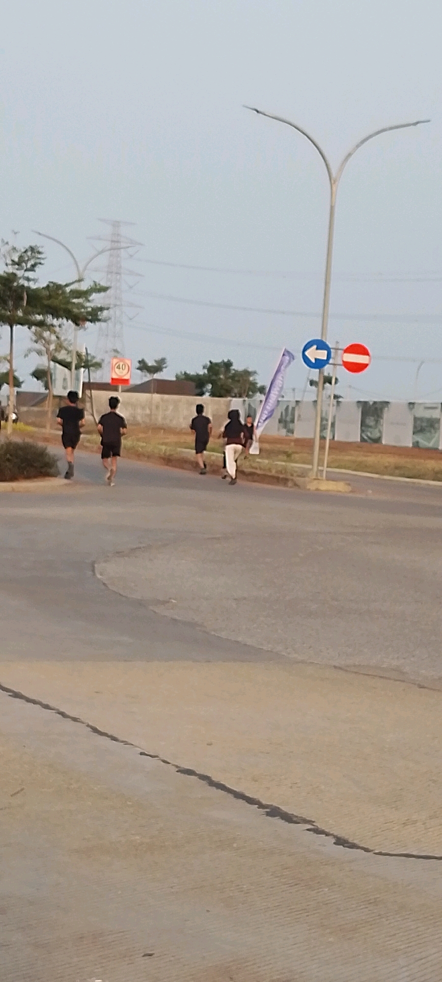 In Satriamekar, Indonesia, a group of five individuals are running on a paved road. Three individuals in black t-shirts and shorts are in the foreground, running from left to right. Two other individuals are further ahead, with one in a white shirt and black pants, and another obscured. A purple and white flag is visible on a pole to the right of the runners. Road signs indicating a speed limit of 40 and a no entry restriction are present. A tall power line tower is visible in the background. The scene takes place in an outdoor urban environment with a light-colored sky.