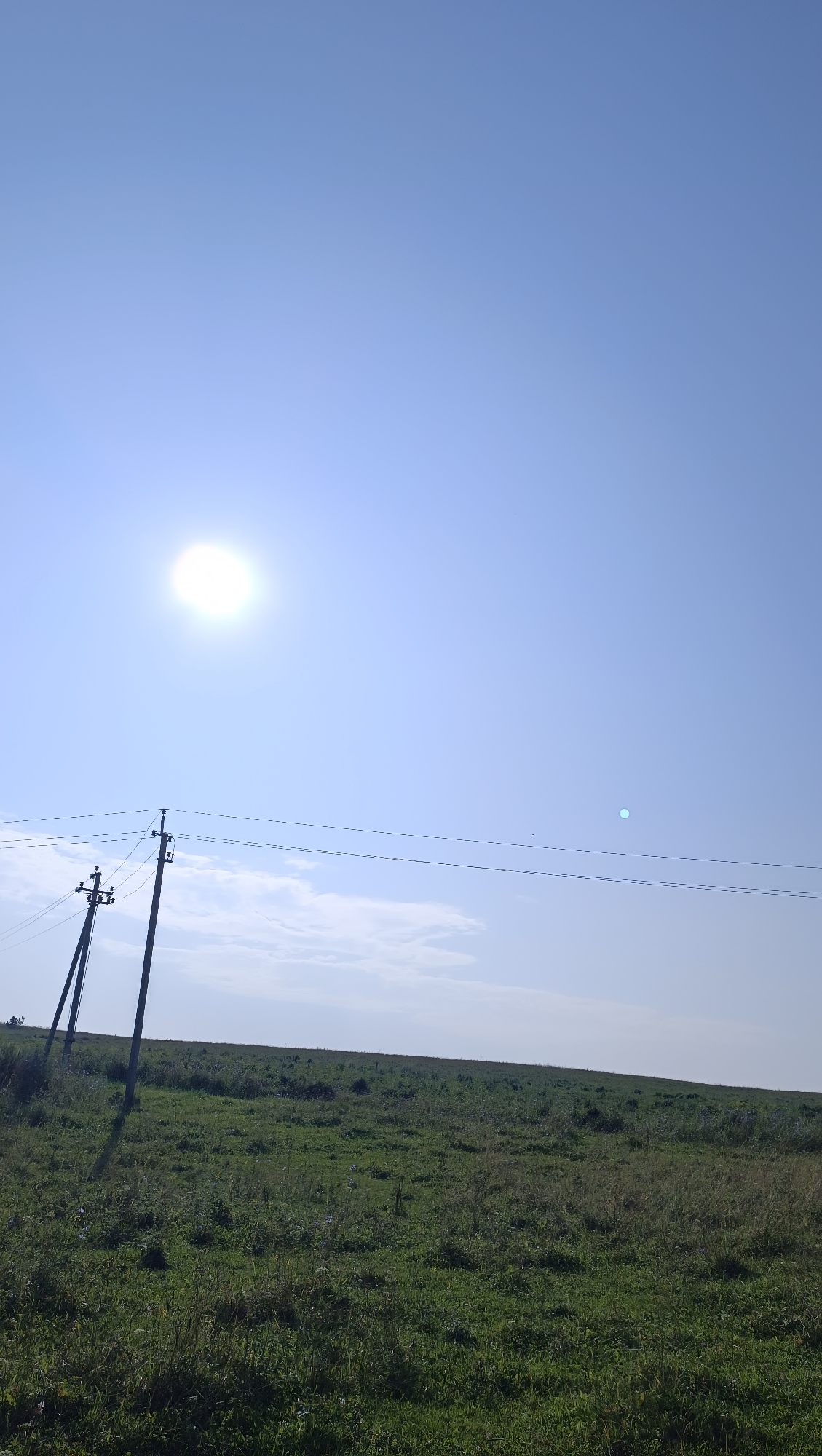 The image depicts an outdoor scene with a bright sun in a clear blue sky. In the foreground, there is a grassy field with some wildflowers. Two telephone poles with wires stretching between them are visible in the middle ground, leaning slightly to the left. In the background, the land slopes upwards, forming a gentle hill. The overall impression is a rural landscape under a sunny sky, possibly in the city of Karsha, Russia, given the context. There are no people or animals visible in the image. The environment appears natural and undeveloped.