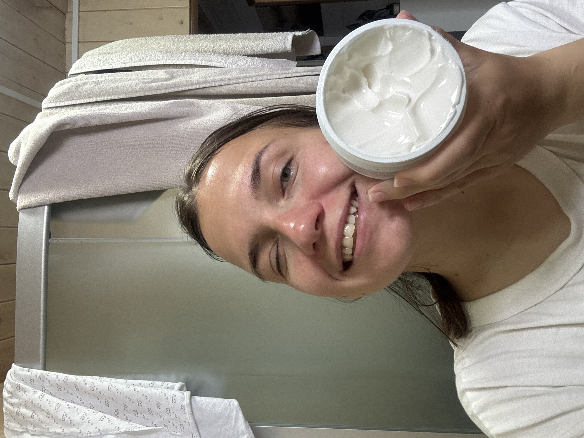 A young woman is depicted in a close-up, rotated image, smiling broadly at the camera. She holds an open white tub filled with a thick, white, creamy substance, likely a cosmetic product such as a moisturizer or body butter, displaying visible swirls and peaks. Her face appears somewhat shiny, and her cheerful expression reveals her teeth in a wide smile. She is wearing a light-colored t-shirt.
The background elements suggest an indoor setting, possibly a bathroom or personal care area. In the upper left, light-colored, folded towels are neatly stacked on a surface, next to wooden paneling. A metallic or silver frame, potentially belonging to a shower enclosure or a mirror, is visible on the left side of the frame. Towards the bottom, a patterned towel can be seen with repeated designs that clearly include the number "13". The woman appears to be engaged in a personal skincare routine. No specific time of day or external weather conditions are discernible. The location context for this media is Раздолье, Russia.