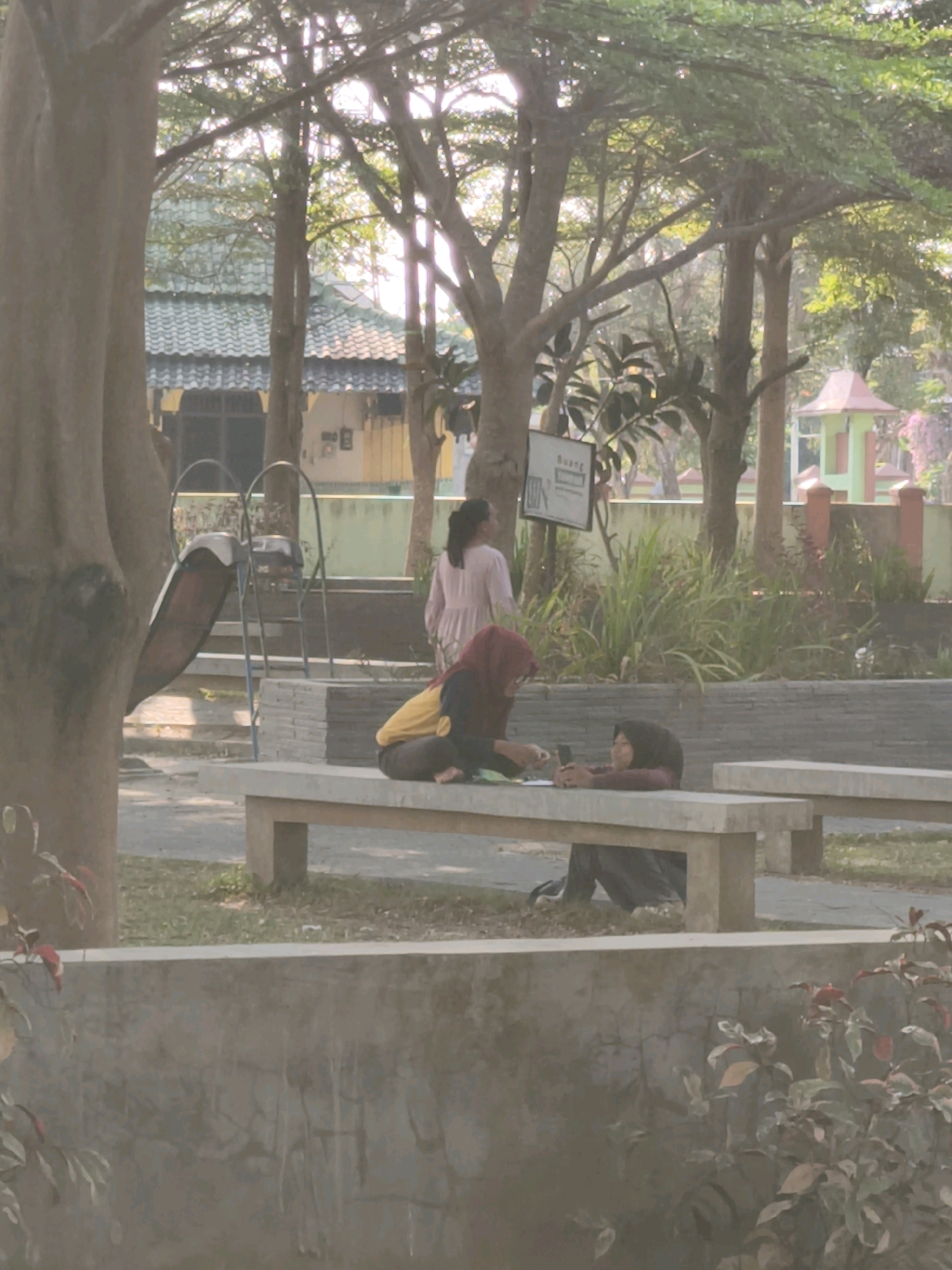In Desa Dibal, Indonesia, a park scene unfolds under dappled sunlight filtering through trees. Two women are seated on a concrete bench. The woman on the left, wearing a red hijab and a yellow and black top, is engaged in an activity on her phone. The woman on the right, wearing a maroon head covering and a maroon top, is also looking at her phone. Behind them, a playground slide is visible, suggesting a recreational area. The environment features lush greenery, trees, and other park amenities. A building with a distinctive green tiled roof is situated in the background, adding to the Indonesian architectural context of the location.