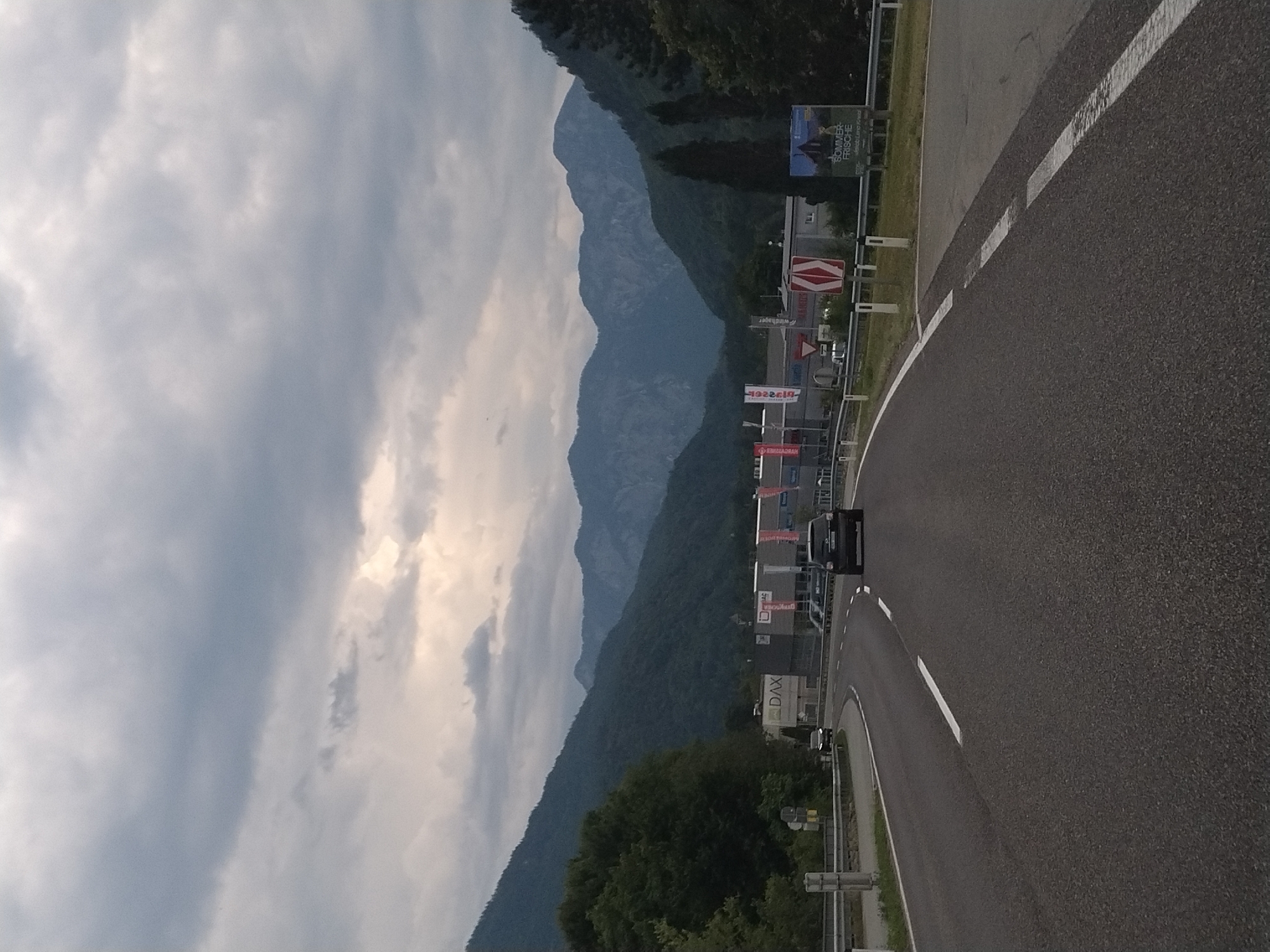 This is a photograph taken from a vehicle driving on a two-lane road in the municipality of Bad Ischl, Austria. The road curves to the right, leading towards a line of commercial buildings on the left side of the image and a billboard on the right. In the background, a range of verdant, mist-shrouded mountains dominates the horizon under a cloudy sky. The sky is a mix of dark grey and lighter, broken clouds, with a faint area of light peeking through in the upper center.
On the left side of the road, a series of commercial structures are visible, including one with the prominent "DAX" logo. There are flags and signage for various businesses. A black SUV is driving ahead on the road, and another dark-colored vehicle is parked or moving slowly near the buildings. The road itself is asphalt, marked with a solid white line on the left and dashed white lines in the lane. A metal guardrail runs along the side of the road.
On the right side of the road, a billboard advertising "SOMMER FRISCHE" (Summer Freshness) is visible. Two diamond-shaped warning signs with red and white chevron patterns are also present. The environment is characterized by rolling hills and mountains covered in dense forest. The overall impression is of a scenic drive through a semi-urban area nestled within a mountainous landscape. The time of day appears to be overcast afternoon or early evening.