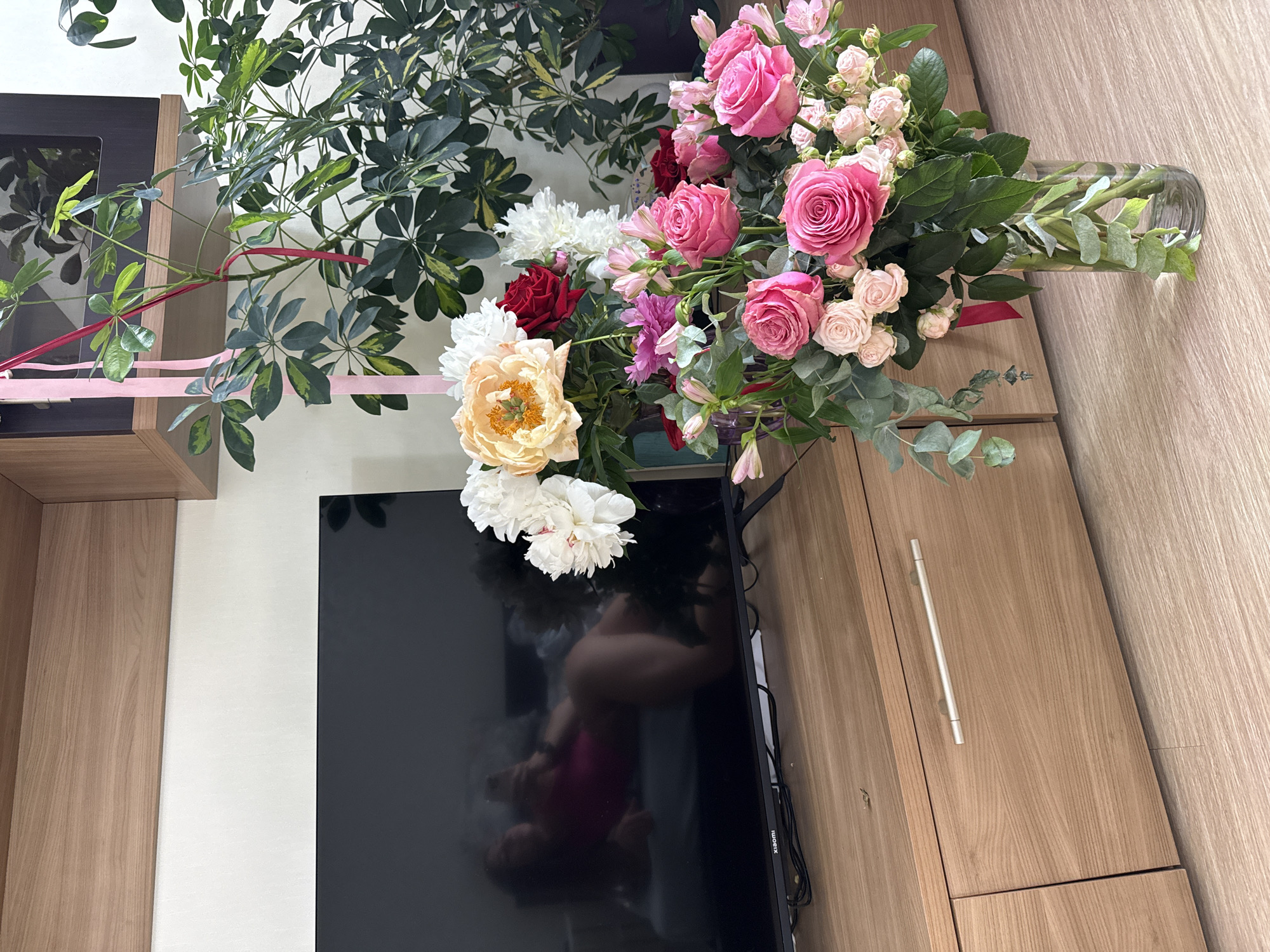 A bouquet of flowers is displayed on top of a television, next to a wooden cabinet. The bouquet features a variety of flowers including pink roses of different sizes, white peonies, a yellow peony, red roses, and some smaller pink and white flowers. Green foliage, including eucalyptus, is mixed throughout the arrangement. The flowers are in a clear glass vase. Behind the bouquet, a large leafy plant with variegated green and yellow leaves is visible, with pink ribbons tied around some of its branches.
The television screen is black and reflects a person wearing a pink top. To the right of the television is a wooden cabinet with a light wood grain finish and a silver handle. A dark shelf unit is partially visible on the left side of the image. The overall setting appears to be indoors, possibly a living room or entertainment area. There is no indication of the time of day or weather. No visible text is present in the image, apart from a small label on the bottom right of the television which reads "XIAOMI".
