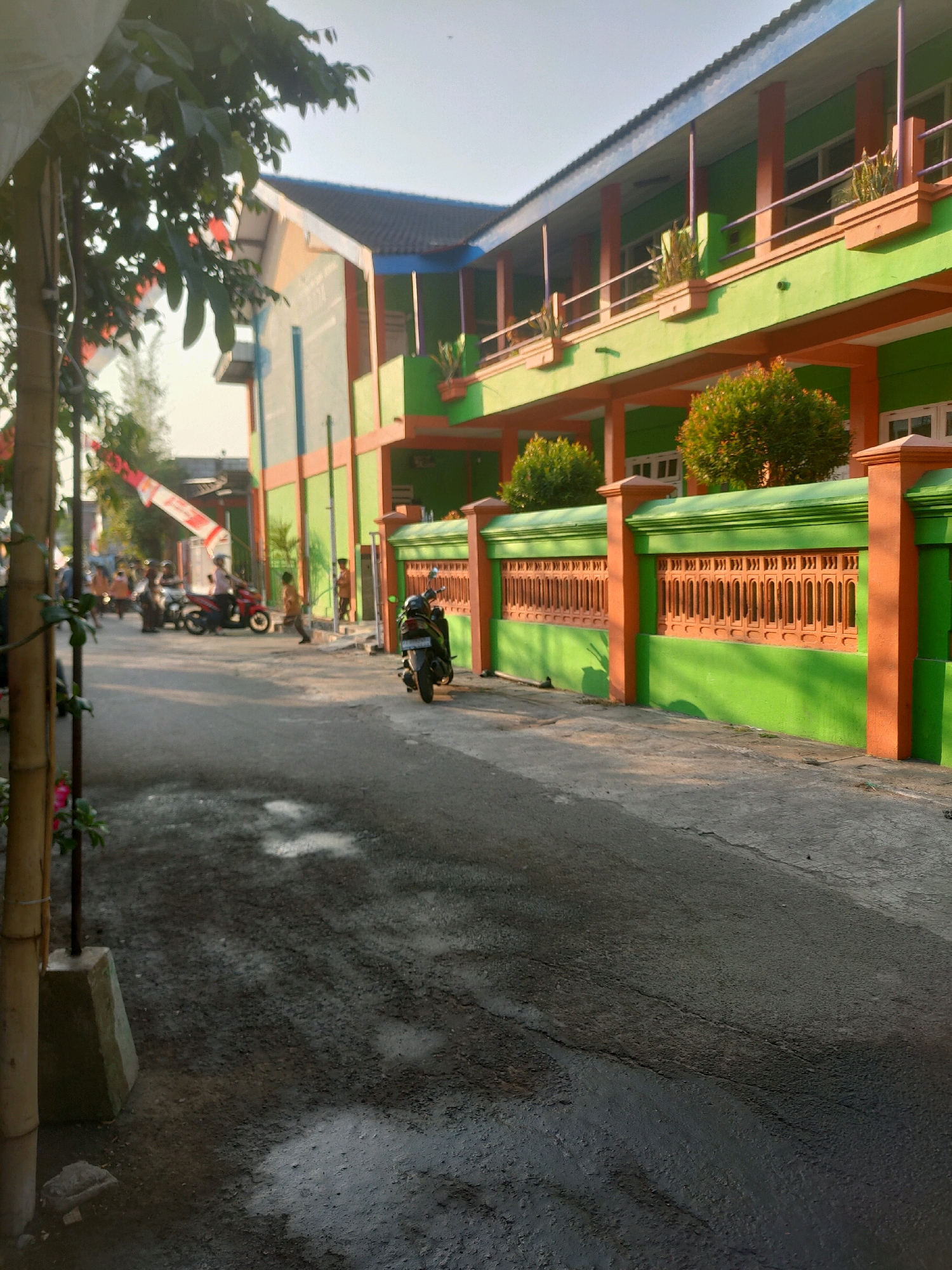 The image depicts a street scene in front of a green and orange building in Nganjuk, Indonesia. The building appears to be a school or similar institution, with multiple stories, balconies, and decorative fencing. A paved road with some wet patches runs in front of the building. Several people are visible, some on motorcycles and others walking. In the foreground, a bamboo structure with leaves is on the left side of the image. In the background, a red and white flag is partially visible. The lighting suggests it is daytime, possibly late afternoon, with sunlight casting shadows.