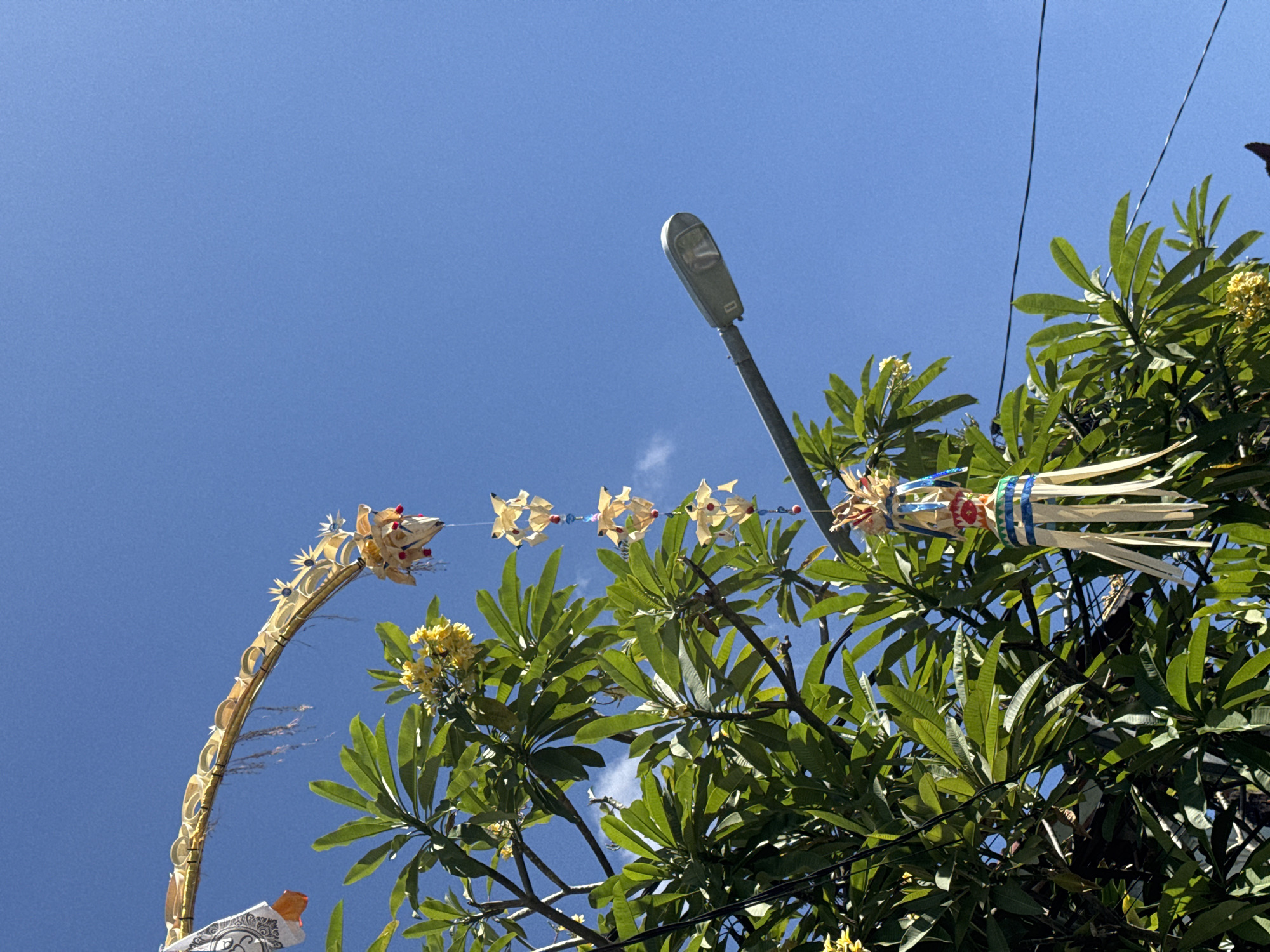 In Kuta, Indonesia, on a clear day with a bright blue sky, a large green leafy tree dominates the left side of the frame, its branches laden with foliage and a few small yellow flowers. Hanging from the branches are intricate Balinese decorations. One is a long, arched golden structure adorned with circular motifs and dangling elements, likely a "penjor," a traditional Balinese offering. Below this, several smaller, delicate decorations made of folded leaves, beads, and red spheres hang in a vertical line. Further down, another decorative element with colorful bands of blue and green, and white streamers, is suspended. In the upper right quadrant, a modern street lamp with a grey arm and housing is angled upwards, its light fixture clearly visible. Thin black electrical wires stretch across the sky in the middle and upper right portions of the image, suggesting an urban or semi-urban setting. The scene captures a blend of cultural decoration and everyday infrastructure against a backdrop of tropical vegetation and clear skies.