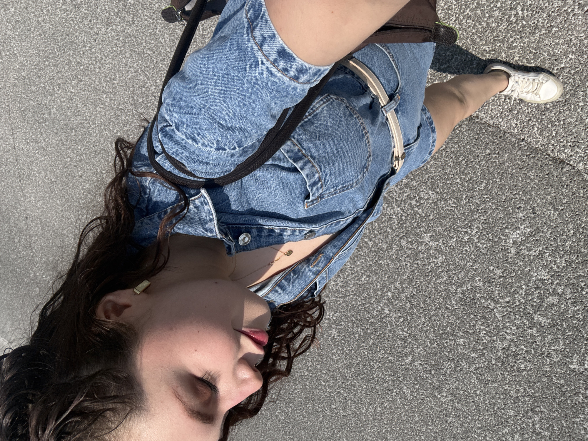 A woman is standing on a gray asphalt surface, likely a street or sidewalk, based on the uniform texture. She is wearing a blue denim dress or jumpsuit, accessorized with a white belt with a metallic buckle, and white sneakers. The woman also sports a gold necklace and a simple gold earring. A dark-colored strap, possibly from a bag, is across her chest. The angle is from above, looking down at the woman. She has dark, curly hair, and her eyes are closed. The location cannot be determined to be Mexico City based on the image.