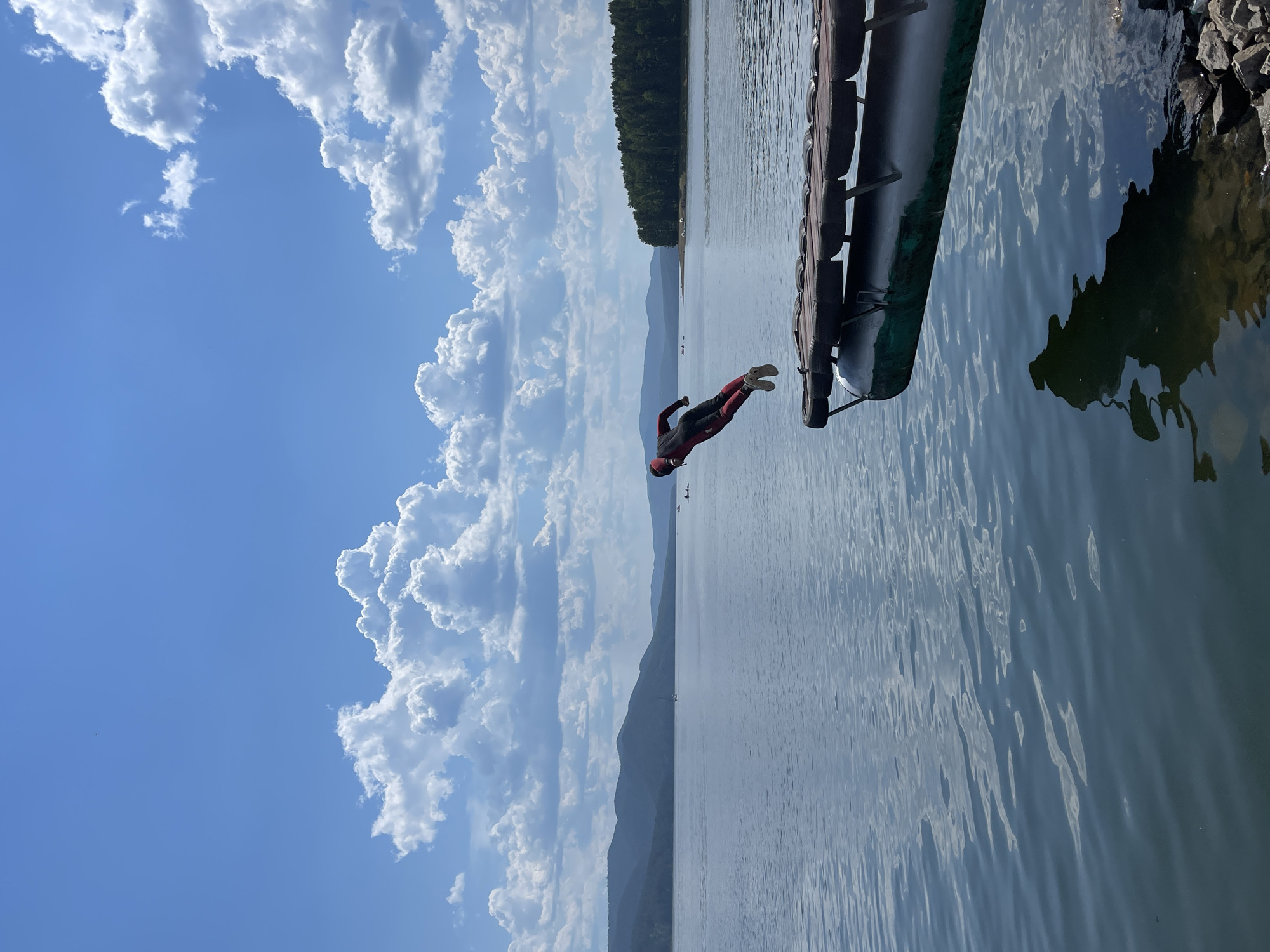 In Divnogorsk, Russia, a person wearing a red and black wetsuit is captured mid-air as they jump from a wooden dock over a calm body of water. The dock, made of wood planks supported by what appears to be a green cylindrical float and tire fenders, extends into the frame from the right. The person is positioned over the water, with their body contorted in a dynamic pose, suggesting they are in the process of diving. The background features a vast expanse of water, a tree-lined distant shore with rolling hills, and a bright blue sky filled with fluffy white clouds. The water reflects the sky and surrounding landscape, creating a serene atmosphere. Several small boats are visible in the distance on the water. The overall scene depicts an active moment in a picturesque natural setting.