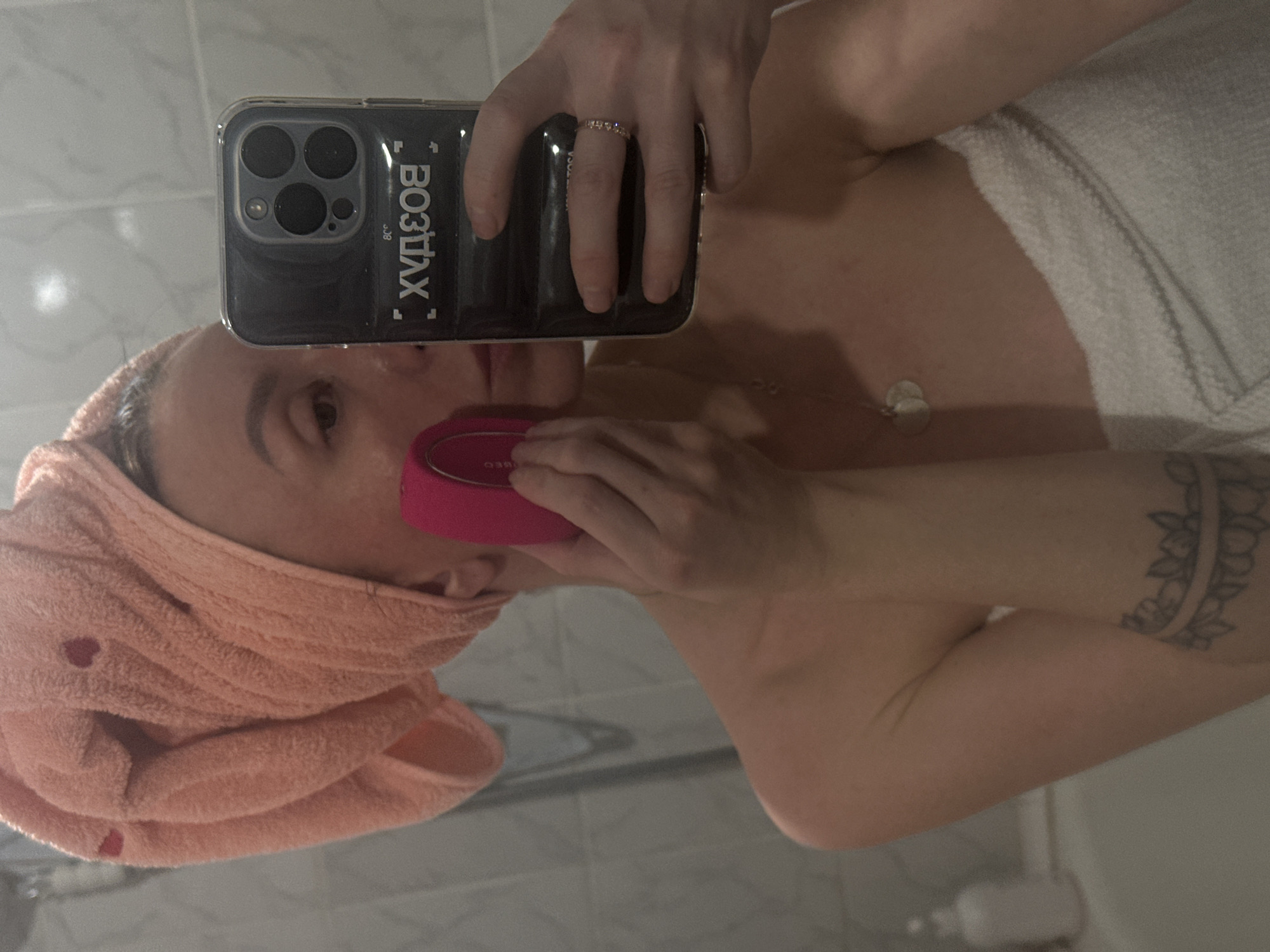 A person is captured in a mirror selfie, seen from the chest up, engaged in a skincare routine. Their head is wrapped in a peach-colored towel adorned with small heart patterns, and a white towel is draped around their body, suggesting a post-shower or bath setting. They are holding a pink, oval-shaped facial cleansing device (identifiable as "FOREO" from visible text) to the right side of their face.
The person is holding an iPhone with a triple camera array in a clear case, visible as the source of the reflection. On the phone case, reversed text "ВОЗДУХ" (VOZDUKH, meaning "AIR" in Russian) and smaller text "338" or "JOB" are visible. A gold-colored ring with small stones is on the ring finger of the hand holding the phone. A silver chain with a circular pendant hangs around their neck.
A tattoo, appearing as a band of floral or leafy design, is visible on their upper right arm (left arm in the mirror reflection). The background shows light-colored square wall tiles, indicative of a bathroom. The overall lighting is consistent with indoor artificial light. The image is oriented sideways.