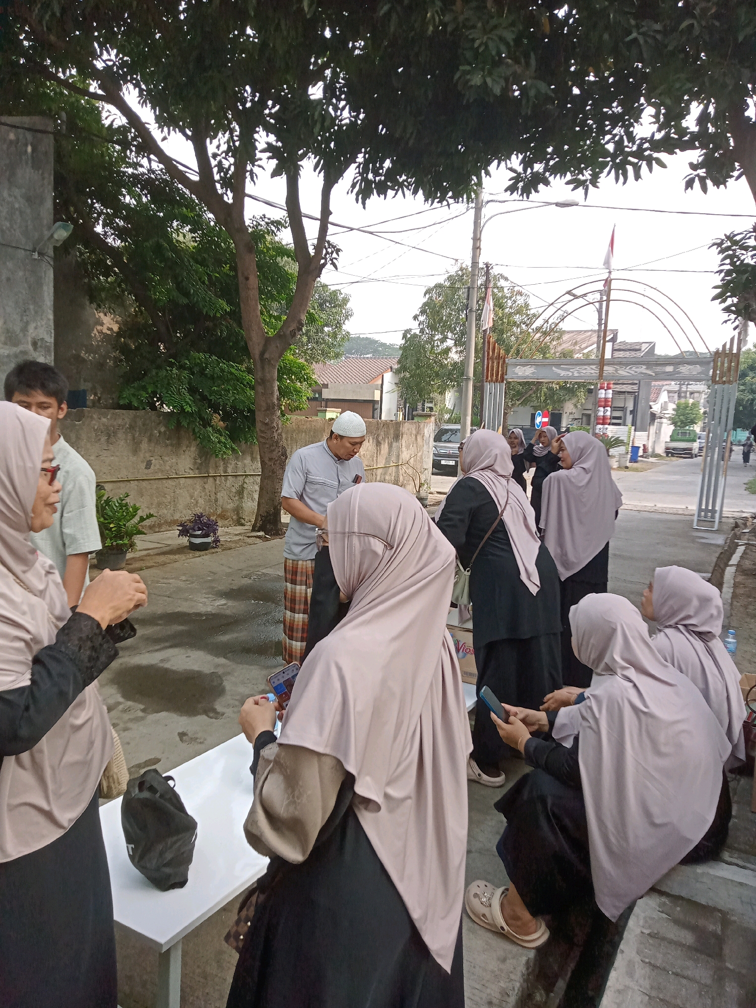In Karawang, Indonesia, an outdoor scene depicts a group of people gathered near a white table. Three individuals, two women and one man, are prominent in the foreground. A woman on the left, wearing a beige hijab and a black garment, is holding a small object, possibly a phone, in her hands. To her right, a man in a light blue shirt and a beige and white checkered sarong is looking down. Another woman, also in a beige hijab and black attire, is seated on the ground to the right, looking at her phone. Behind them, several other individuals, predominantly women in hijabs, are standing or walking, some looking towards a decorative arched gate in the background. The environment includes a concrete wall, trees with green foliage, a street, and houses. The overall atmosphere suggests a casual gathering or event.