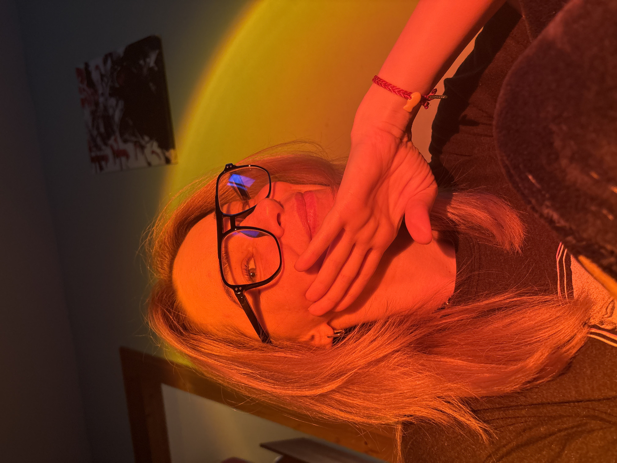 The image displays a close-up portrait of a person, likely female, with long, reddish-auburn hair. They are wearing black-framed glasses, and one eye is visible, looking towards the viewer. A hand covers their mouth and lower face, with the fingers gently resting on their cheek and chin. On the wrist of the covering hand, a red braided bracelet adorned with a small yellow charm, possibly shaped like a fish or an abstract symbol, is visible. The person is dressed in a dark-colored garment, possibly a top or jacket, parts of which are visible on the right side of the frame.
The setting is indoors, characterized by striking, artificial lighting. A dominant warm glow, transitioning from vibrant yellow to orange and deep red, illuminates the wall behind the subject and casts rich colors across their hair, face, and arm. This light creates a gradient effect, suggestive of a sunset lamp or similar light source. The surrounding areas of the room are dimly lit, indicating a low-light environment or nighttime. In the background on the left, a framed abstract artwork with dark and lighter elements hangs on the wall. A wooden frame or the edge of a piece of furniture is also faintly discernible in the lower left.
The activity depicted is a posed portrait. The hand covering the mouth could suggest thoughtfulness, introspection, or a deliberate artistic pose. The subject's direct gaze through their glasses conveys engagement with the camera. The overall mood is intimate and contemplative, strongly influenced by the dramatic, warm lighting. There is no visible text within the image. The location context provided is Unknown, Russia.
