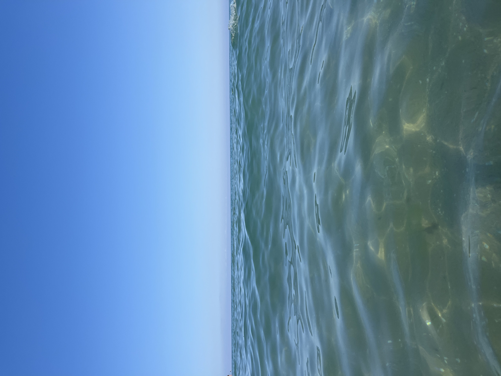 This is an outdoor, vertical, low-angle, eye-level, close-up, full shot of clear, blue sky meeting the ocean with a slight wave. The sky is a gradient of bright blue at the bottom fading to a lighter blue at the top. The ocean is clear and appears shallow, with sunlight glinting off the wavy surface. The water is a light green-blue, and the bottom is visible, with sand and possibly some small rocks or debris. There are some lighter, circular patterns on the seabed, possibly from light refraction or underwater objects. A small wave breaks at the top left of the frame, creating some white foam. The image depicts a peaceful and serene scene, likely during the daytime on a clear day. No people, objects, or text are clearly visible. The image conveys a sense of tranquility and natural beauty. The location context of Emona, Bulgaria suggests a coastal environment.