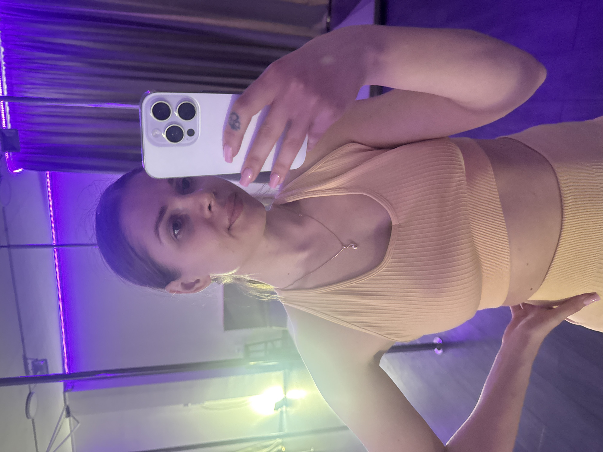 A young woman is depicted taking a mirror selfie with a white smartphone, which she holds with her left hand, obscuring part of her face. She has dark hair pulled back and a neutral expression, looking directly at the phone's camera. She is wearing a ribbed, light peach or beige athletic two-piece set, comprising a sports bra or crop top and matching high-waisted bottoms. A delicate silver necklace with a small, curved pendant is visible around her neck. Her fingernails are painted a light pink, and a small, dark tattoo is visible on the inner side of her right index finger. Her right hand rests near her waist or hip.
The setting appears to be an indoor space, possibly a gym or fitness studio, marked by striking purple ambient lighting from LED strips, casting a distinct glow on the background walls. Reflections of other light sources, some appearing yellowish-green, are also visible. The floor is dark. The image conveys a modern and active atmosphere. The photograph was taken in Batumi, Georgia.