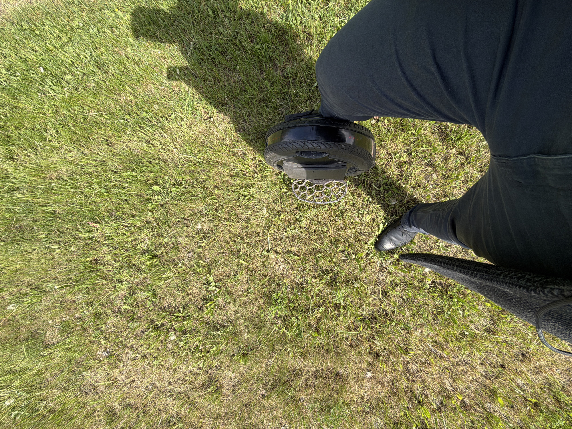 The image depicts a person standing on an electric unicycle on a grassy area. The person is wearing dark pants and black shoes. The unicycle has a black tire and a silver footrest. The background is green grass with some brown patches. The person's shadow is visible on the grass. The location is unknown.