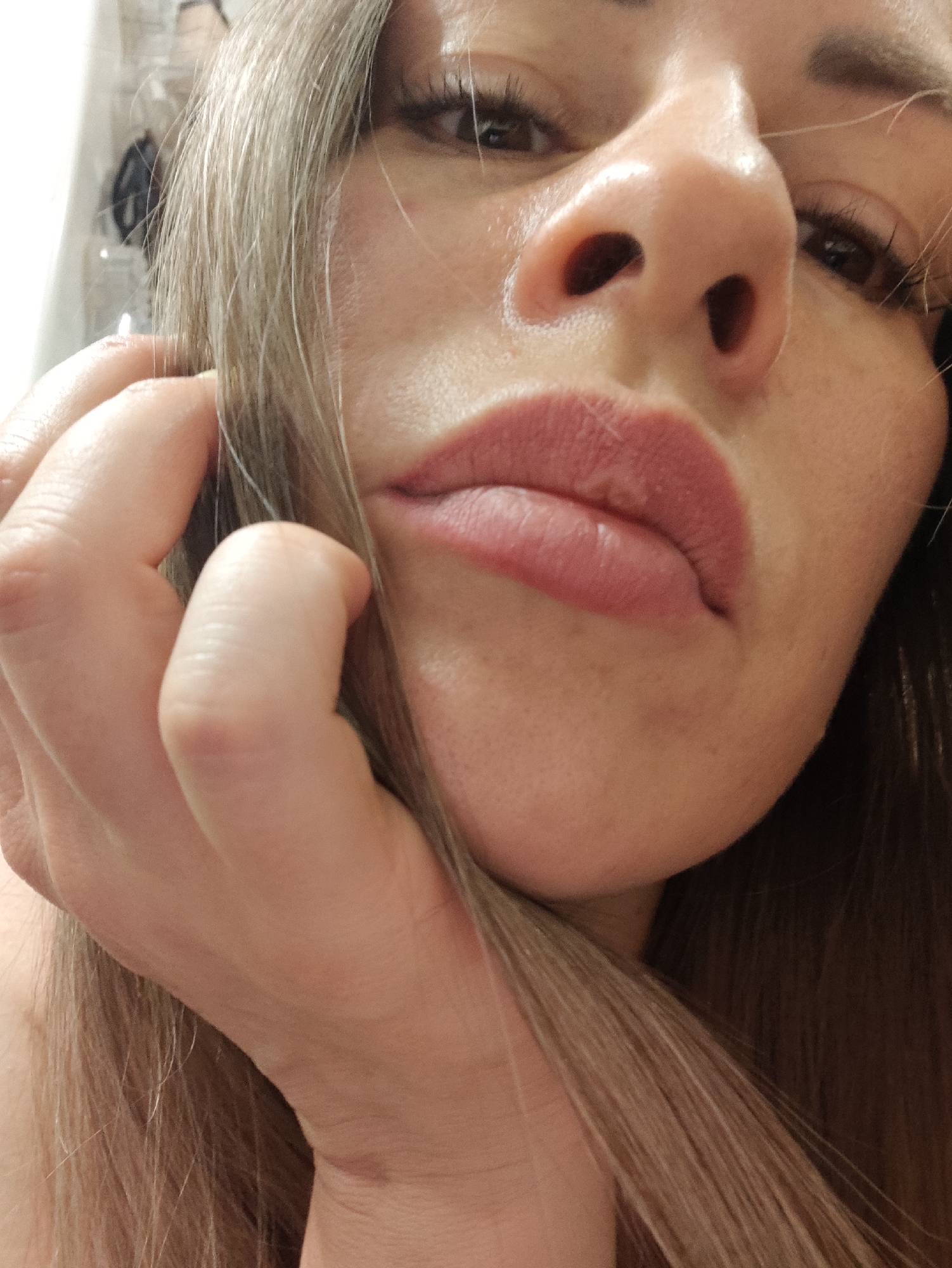 The image is a close-up, low-angle selfie of a woman. Her face is positioned very close to the camera, with her chin and lower nose prominent. She has blonde hair that partially obscures her face. Her hand is raised to her face, with fingers touching her hair and cheek. Her lips are visible and appear to be wearing lipstick. The background is blurred and appears to be an indoor environment, possibly a bathroom due to the reflective surface visible on the left side. There is no information in the image that indicates the location in Almaty, Kazakhstan.
