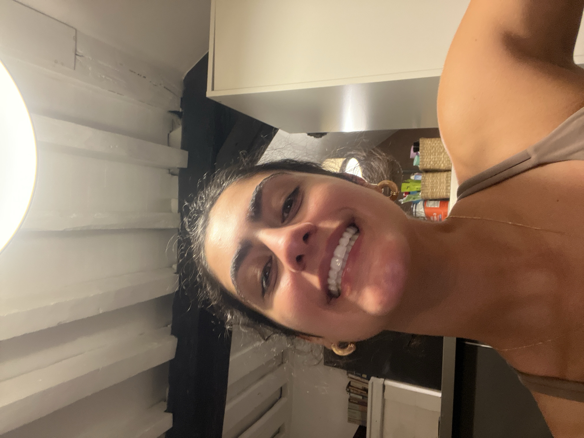 A young woman is shown in a selfie, smiling. She is wearing hoop earrings and a brown tank top. The background appears to be an apartment in Paris, France. There is a white ceiling with wooden beams, a white cabinet, and a shelf with books and baskets.