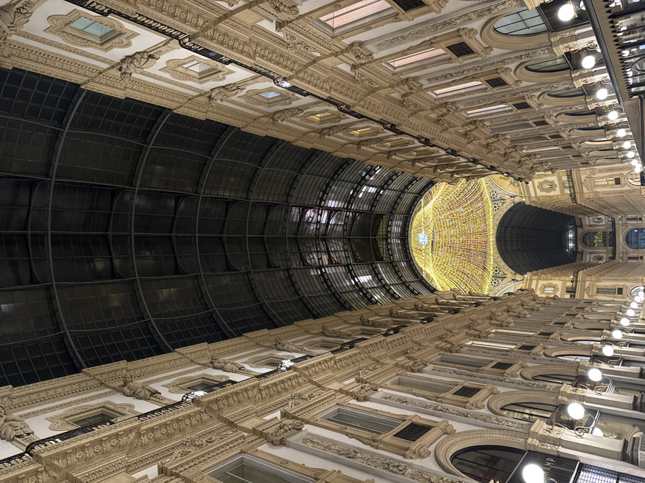 An upward-angle photograph captures the interior of the Galleria Vittorio Emanuele II in Milan, Italy, at night. The scene depicts a grand, covered public passageway flanked by two multi-story, ornately decorated building facades of light-colored stone or stucco, featuring classical architectural elements such as pilasters, balustrades, arched windows, and sculptural details. A vast, dark, arched glass and steel roof spans above the passage. In the distance, where the two main arms of the gallery converge, a large central dome is prominently illuminated by a dense canopy of warm-toned string lights. Below this dome, another arched passage is visible. The lower levels of both facades are lit by spherical white lanterns and internal shop lighting, with partial signage visible on the left facade. No individuals or discernible actions are present in the image.