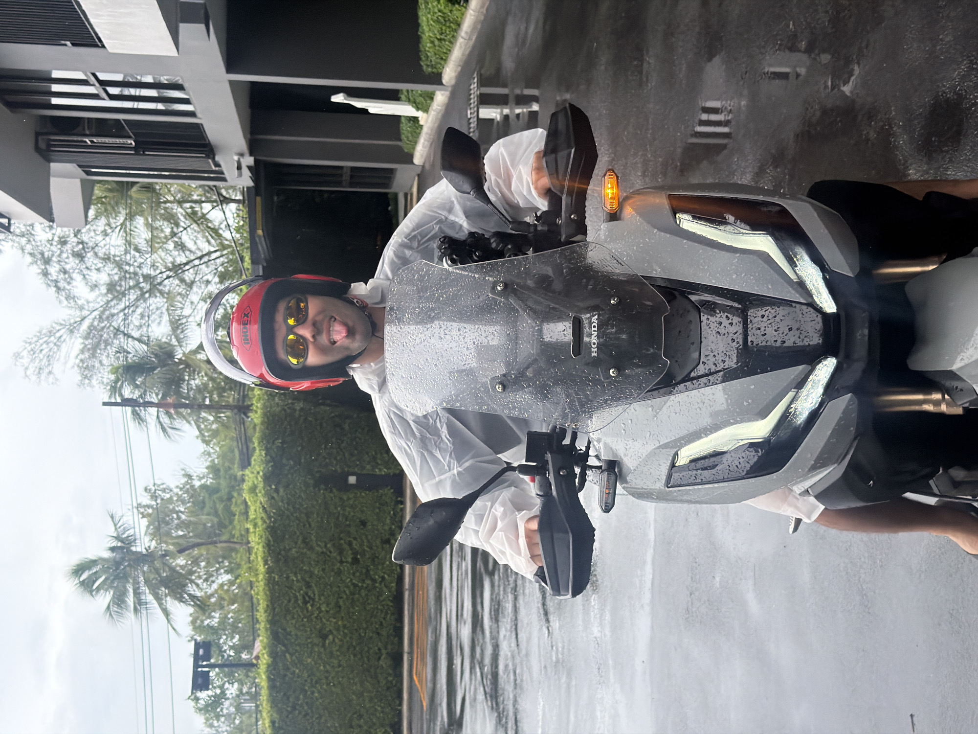 The image depicts a person on a Honda motorcycle in what appears to be Choeng Thale, Thailand, as suggested by the tropical vegetation and architecture. The person is wearing a red helmet with yellow lenses, and a white rain poncho. It is raining, and there are raindrops on the motorcycle's windshield and body. The background includes buildings, trees, and a wet road. The person is sticking out their tongue.