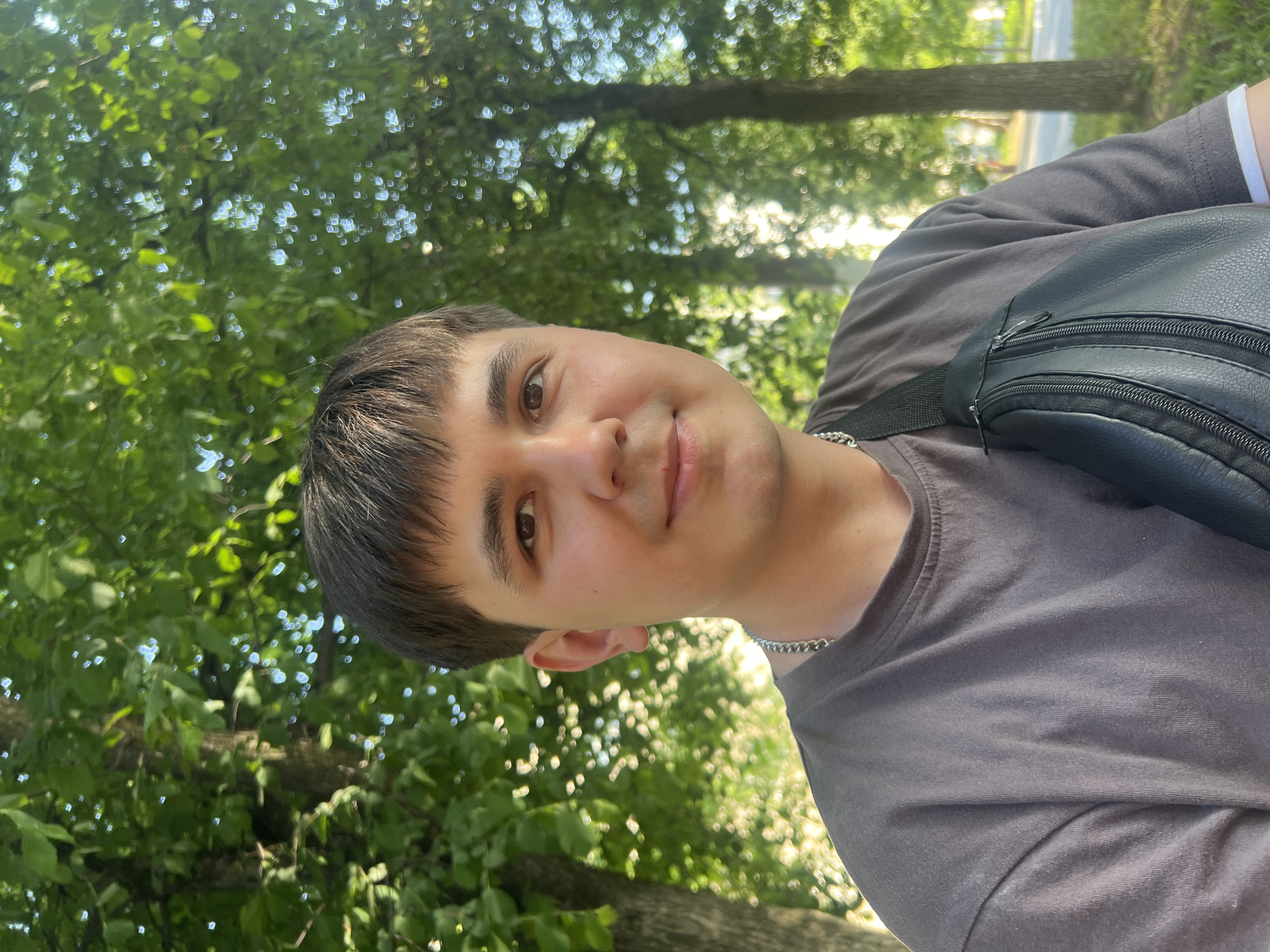 A young man is photographed in an outdoor setting. He has short, dark brown hair and brown eyes, and he is wearing a dark gray t-shirt with a silver chain around his neck. A black shoulder bag is slung over his shoulder. The background shows green foliage, suggesting trees and possibly a park. The location may be in Chaikovsky, Russia, but there is no information in the image to corroborate this claim.