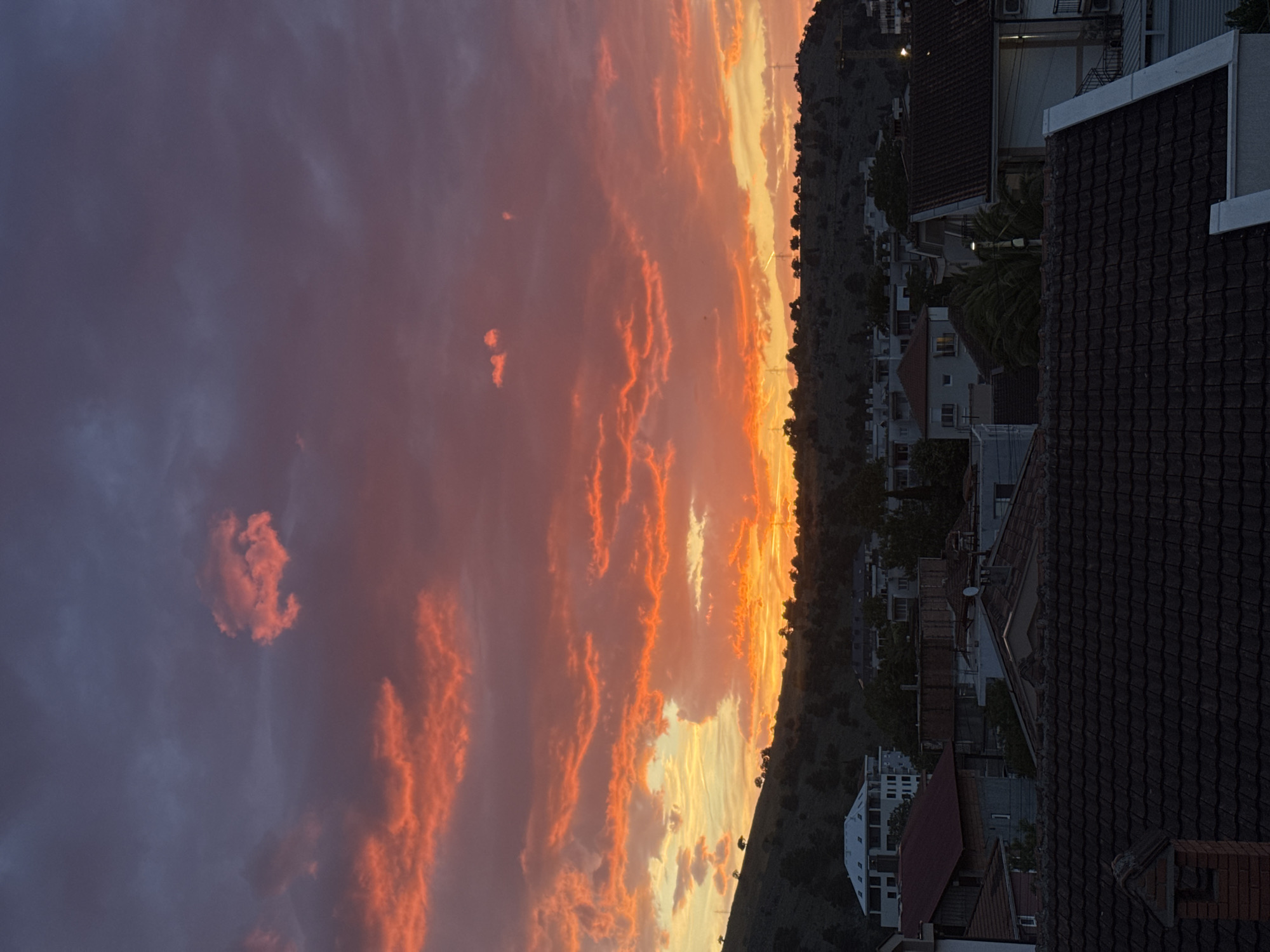 The image captures a sunset over Podgorica, Montenegro, observed from a rooftop perspective. The sky is dramatically colored with hues of orange and pink, reflecting on the clouds. Below the horizon, a tree-covered hill stretches horizontally across the frame, obscuring the landscape. On the right side of the image, rooftops of residential buildings are visible, with their varying heights and roof styles. The foreground is dominated by a dark, tiled roof, providing a visual barrier to the city below. The overall scene suggests a quiet evening in a suburban setting.