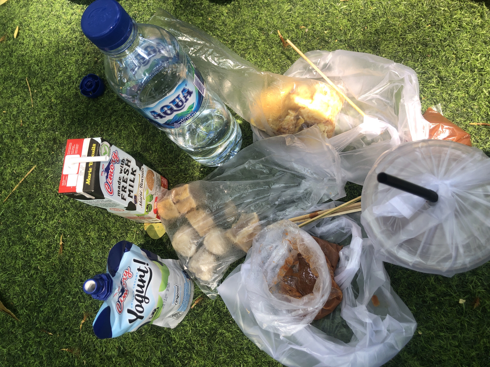 An overhead shot shows a picnic spread out on a green lawn. In the center, a carton of Cimory Fresh Milk rests on the grass. To its left, a pouch of Cimory Yogurt is positioned. To the right, a bottle of Aqua water is open with its cap off, and beside it are several items in plastic bags, including what appear to be food items on skewers and a container of sauce. A blue cap, likely from the water bottle, is also on the grass nearby. The setting appears to be outdoors on a sunny day, with blades of grass forming the background. There are no people visible in the image, and no indication of the city of Pasuruan, Indonesia.