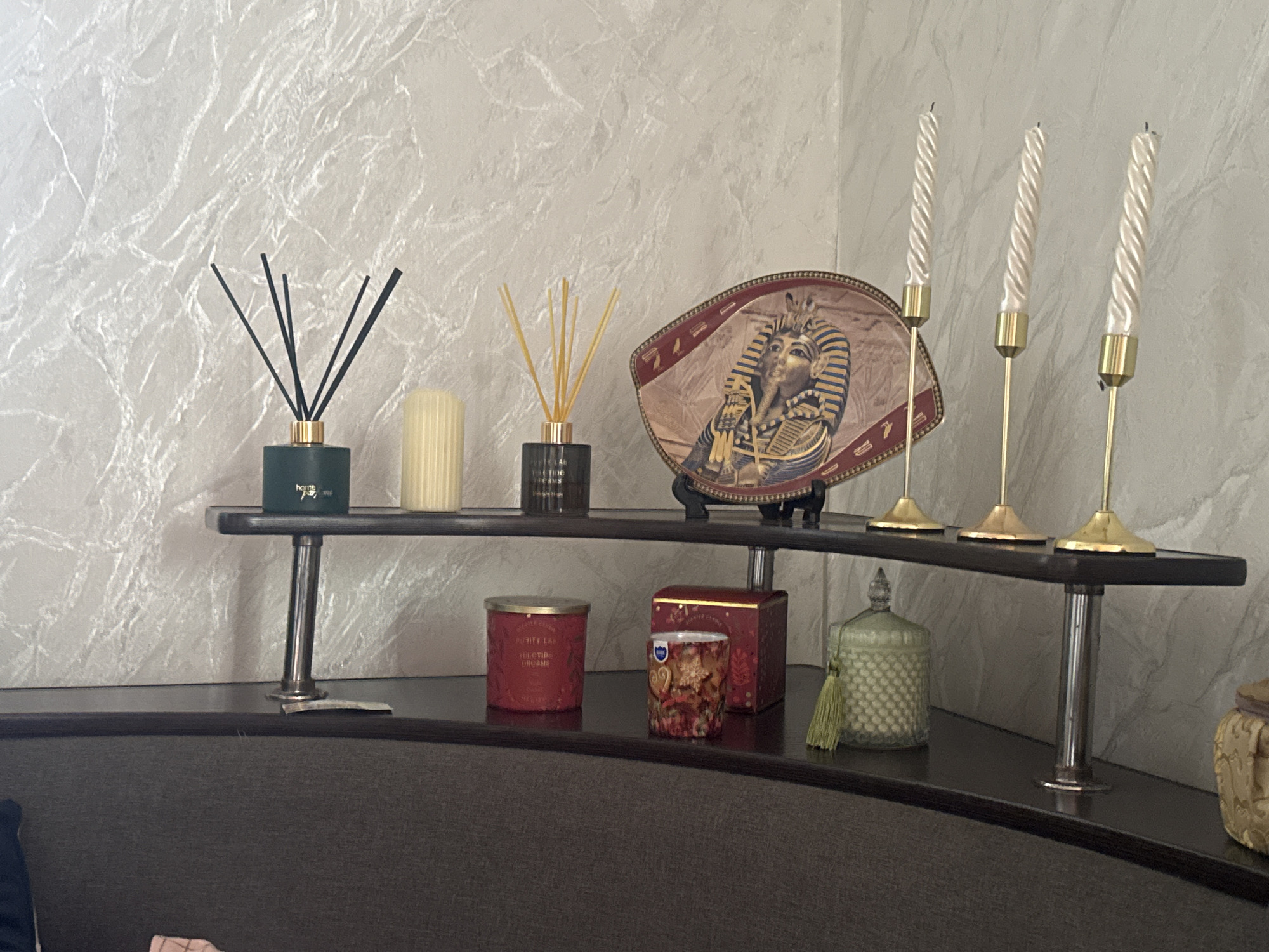 The image depicts a decorative shelf arrangement in what appears to be a residential setting. The shelf is supported by metallic cylindrical legs and displays various objects. On the left, there is a dark green diffuser with black reeds, next to a ribbed, cream-colored candle. Further to the right, a small dark bottle holds yellow reeds. A decorative fan-shaped item featuring an image of King Tutankhamun's mask is positioned behind these items. To the right of the fan are three tall, thin, pearlized candles in gold candle holders.
Below this shelf, on a lower dark surface, are several more decorative items. These include a red cylindrical candle with gold lettering, a smaller candle in a patterned glass holder, and a red and gold rectangular box. To the right of these is a light green glass jar with a textured pattern and a green tassel hanging from it. Partially visible on the far right is another decorative object with fabric textures. The background is a textured, off-white wall. The scene is brightly lit, suggesting indoor lighting. The location is identified as Bohunove, Ukraine. There are no people visible in the image.