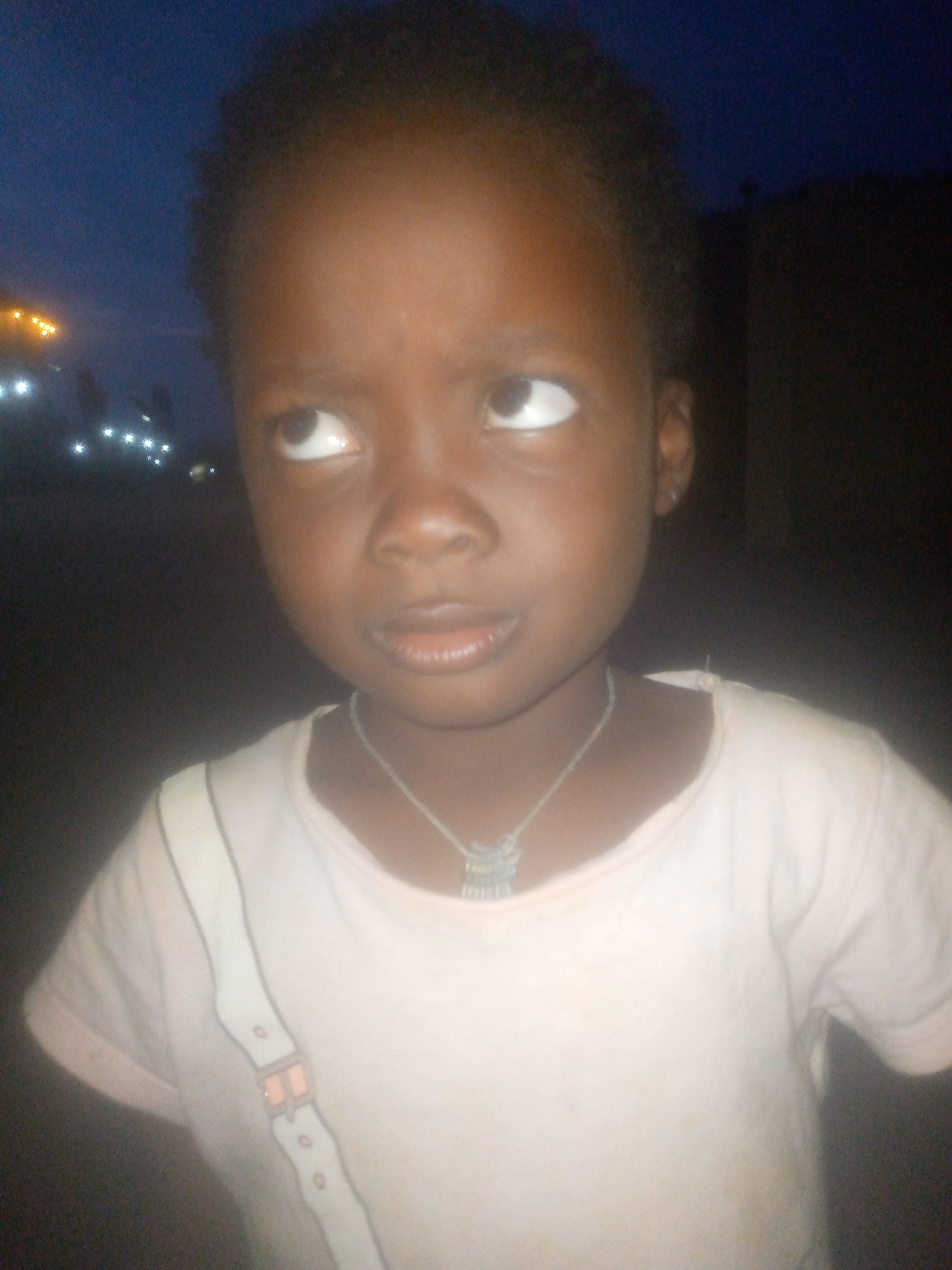 A child with dark skin and dark, close-cropped hair looks upwards and to the left, away from the camera. The child is wearing a light-colored t-shirt with a design of straps and buckles and a necklace with a silver pendant. The setting appears to be outdoors in the city of Ikorodu, Nigeria, at dusk or night. Streetlights are visible in the background, indicating an urban location. A dark building is visible to the right.